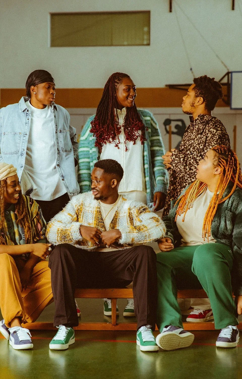 Group of six diverse young adults socializing in a gym or indoor sports space, sitting and standing, engaged in conversation and smiling.