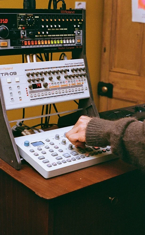 A person operating a music production setup with a Roland TR-09 drum machine and a Roland TR-08 rhythm composer, connected to a computer controller, on a wooden desk in a room with a yellow wall.