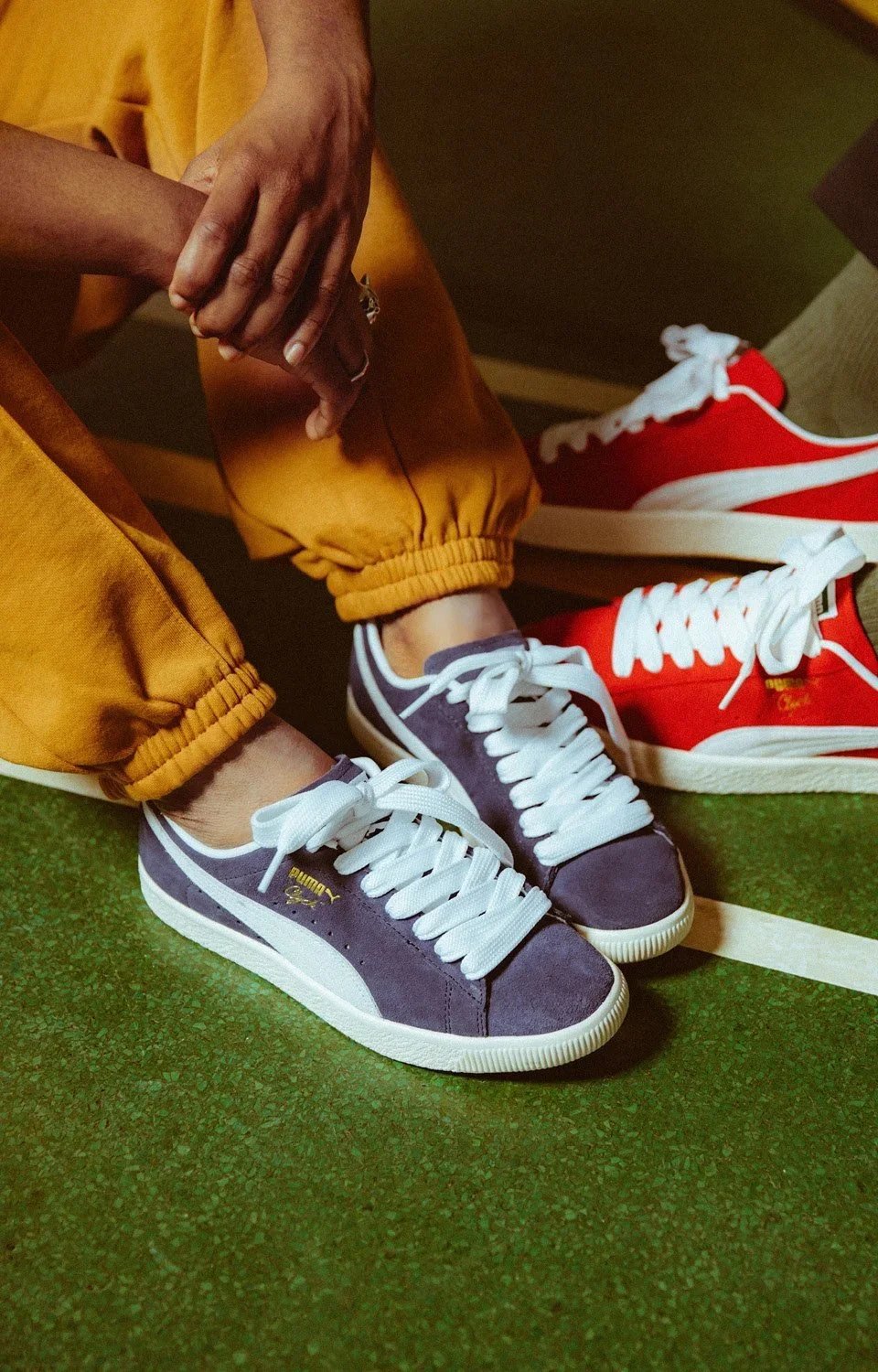 Close-up of a person wearing gray Puma sneakers with white laces and orange pants, standing on a green indoor surface, with another person wearing red sneakers nearby.