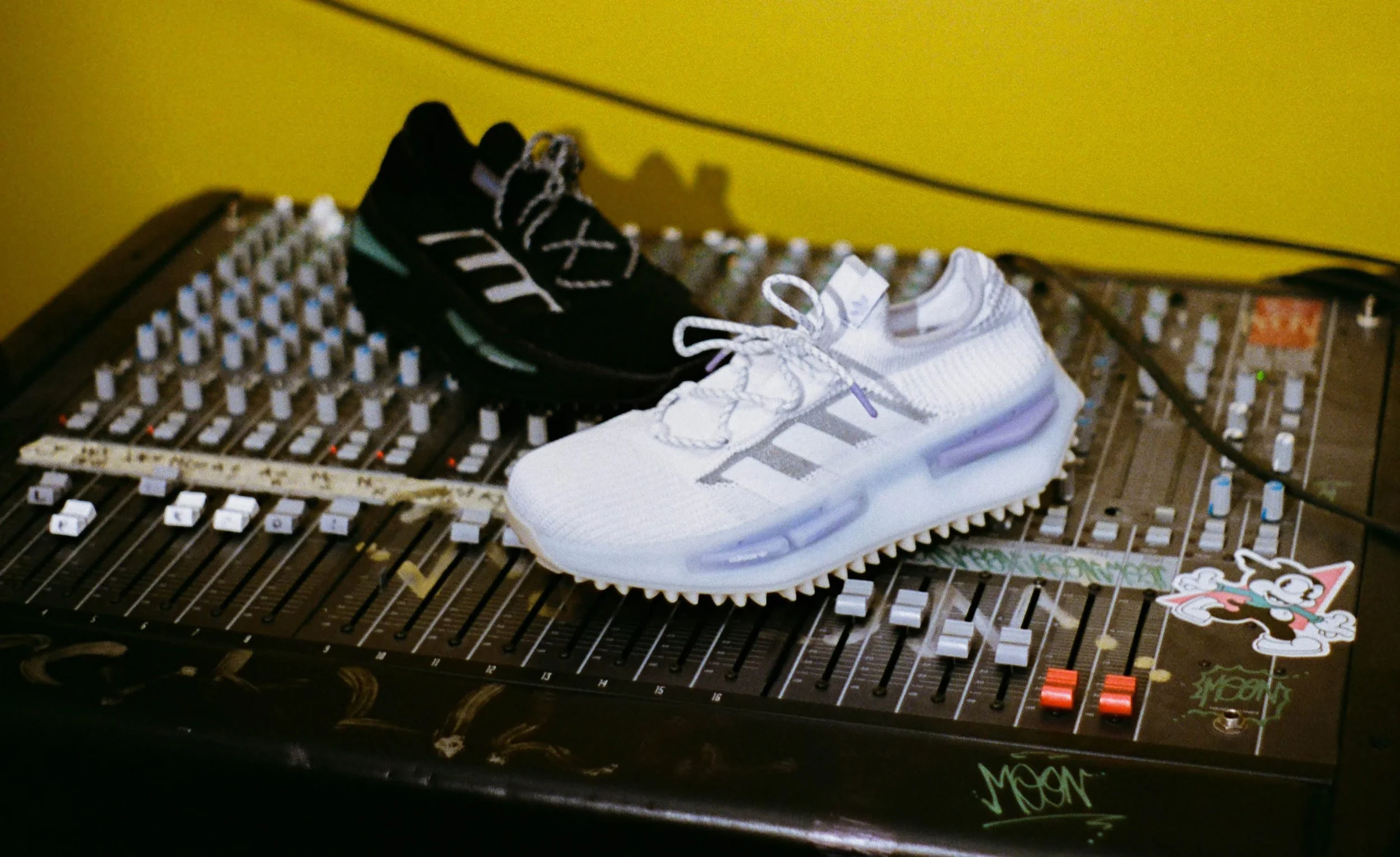 A pair of black and white sneakers placed on a sound mixing console with stickers and colored knobs, against a yellow wall.