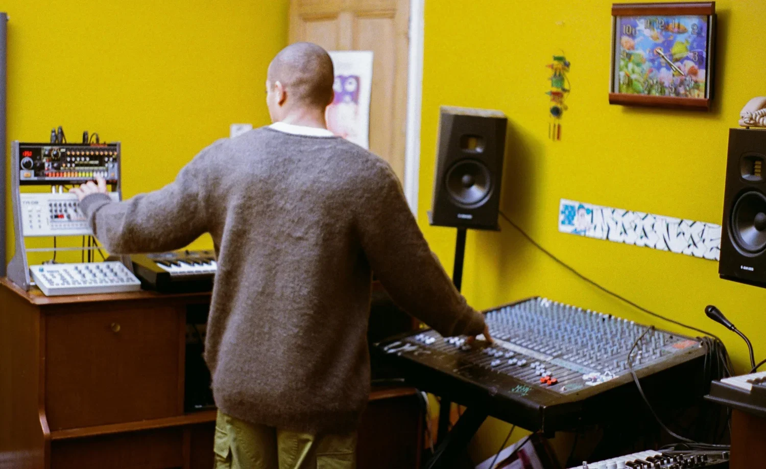 Person operating music production equipment in a recording studio with bright yellow walls, speakers, and a colorful framed picture.