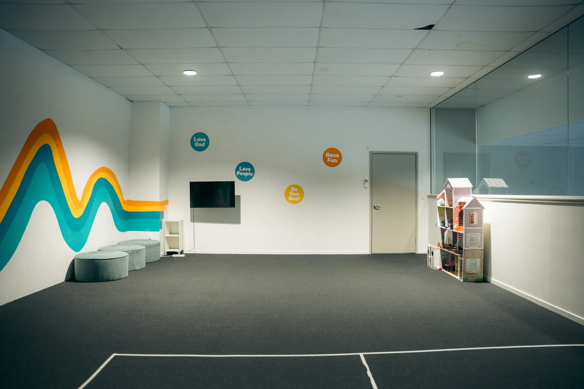 An empty indoor room with a dark carpeted floor, colorful wavy wall art, gray round seating in Caloundra