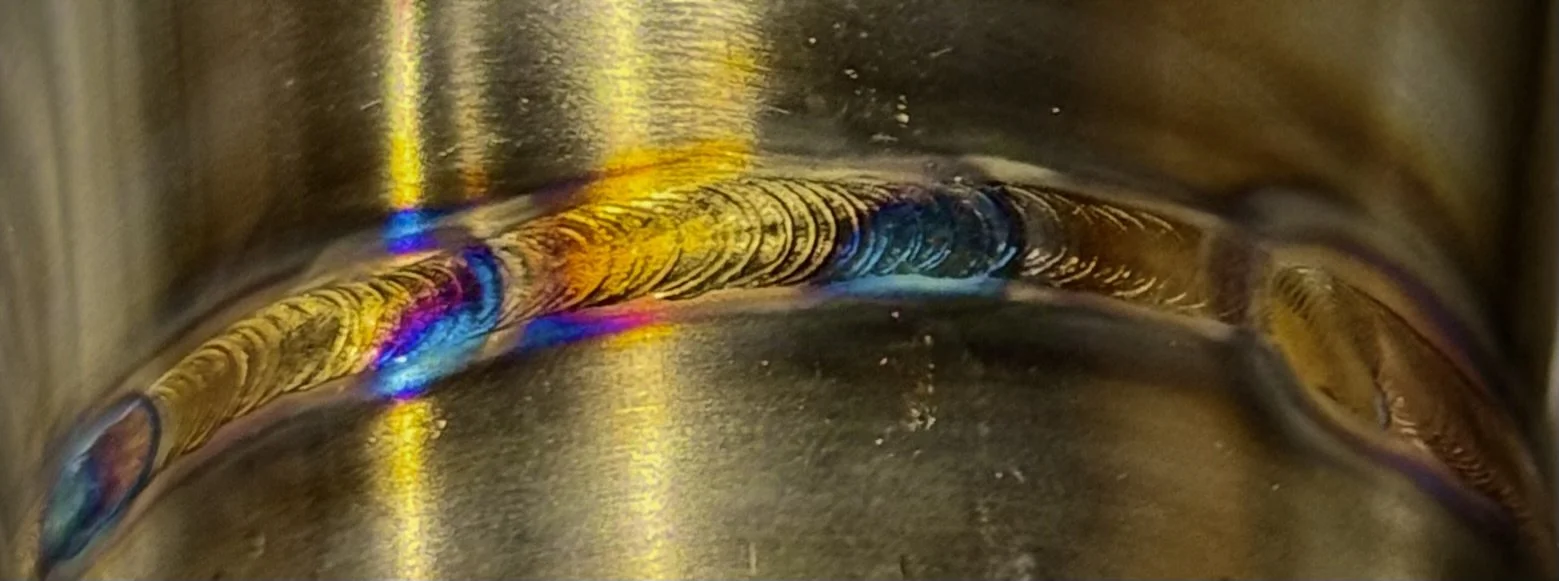 Close-up of a metal pipe with rainbow-colored weld patterns along the curved joint.