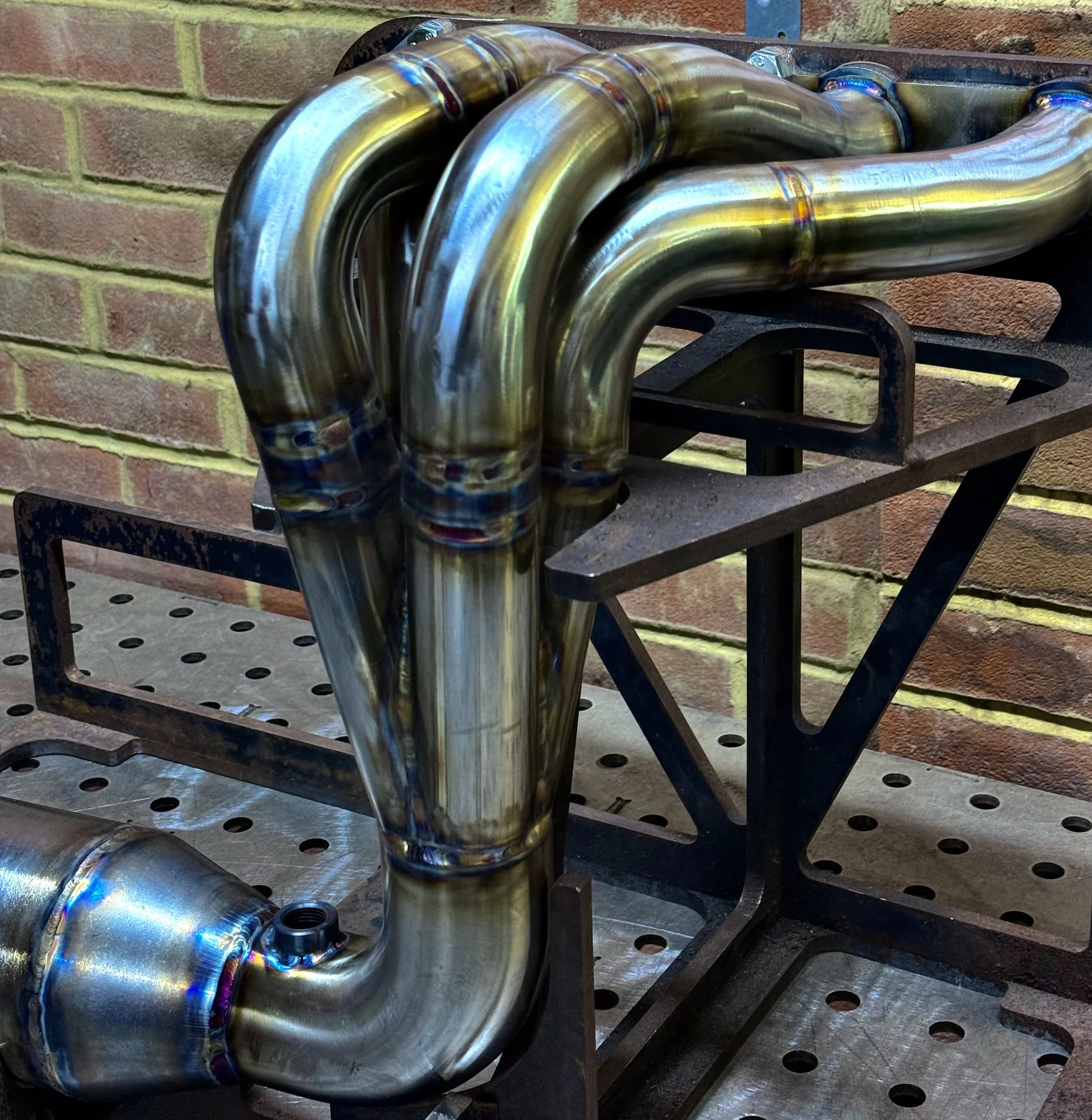 A shiny metal exhaust header pipe with multiple curved sections, mounted on a metal workbench against a brick wall.