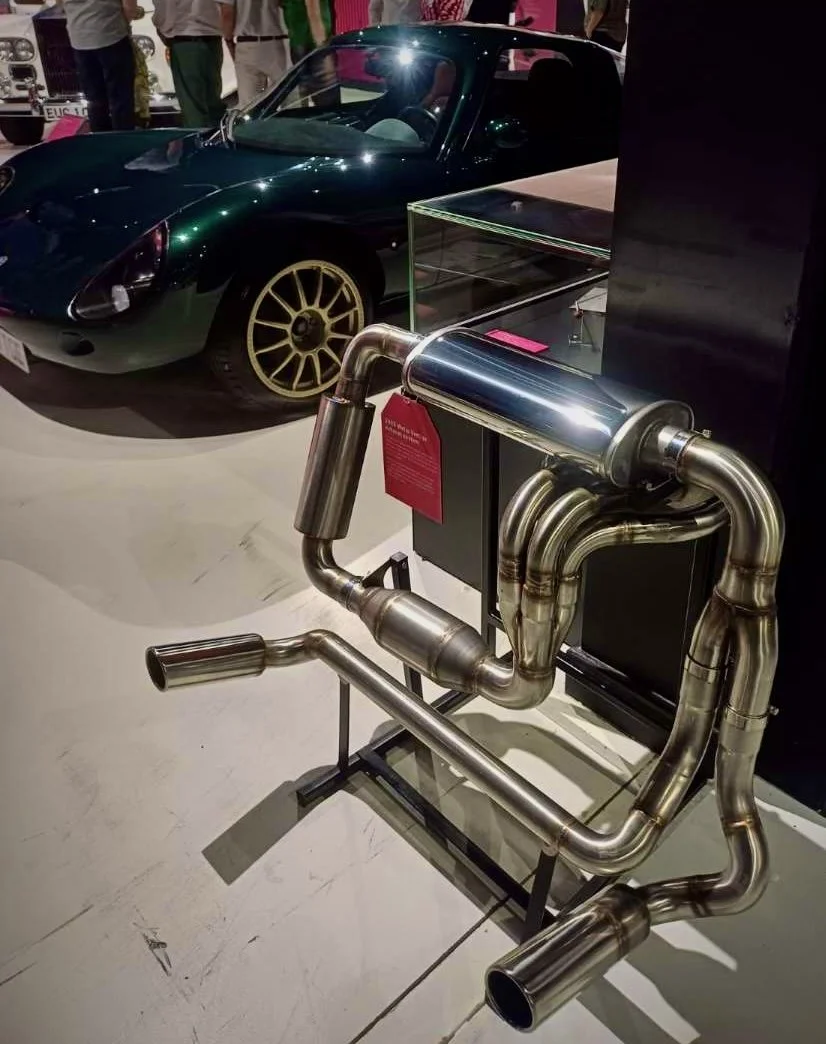 A sleek black sports car with gold wheels displayed behind a stainless steel exhaust system in a museum or exhibition setting.