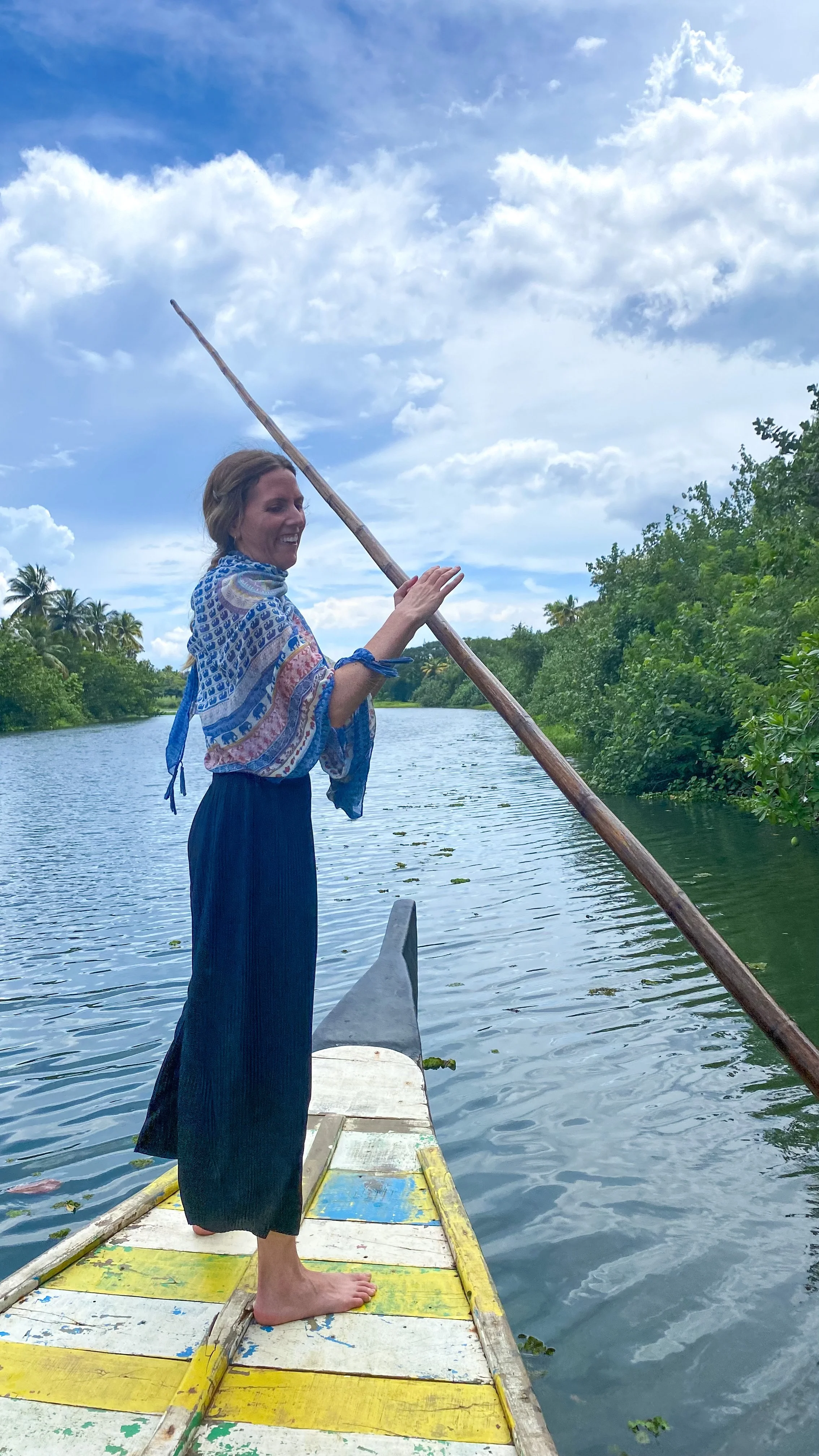 A woman standing barefoot on a colorful boat, paddling on a river surrounded by green trees, with a cloudy sky overhead.