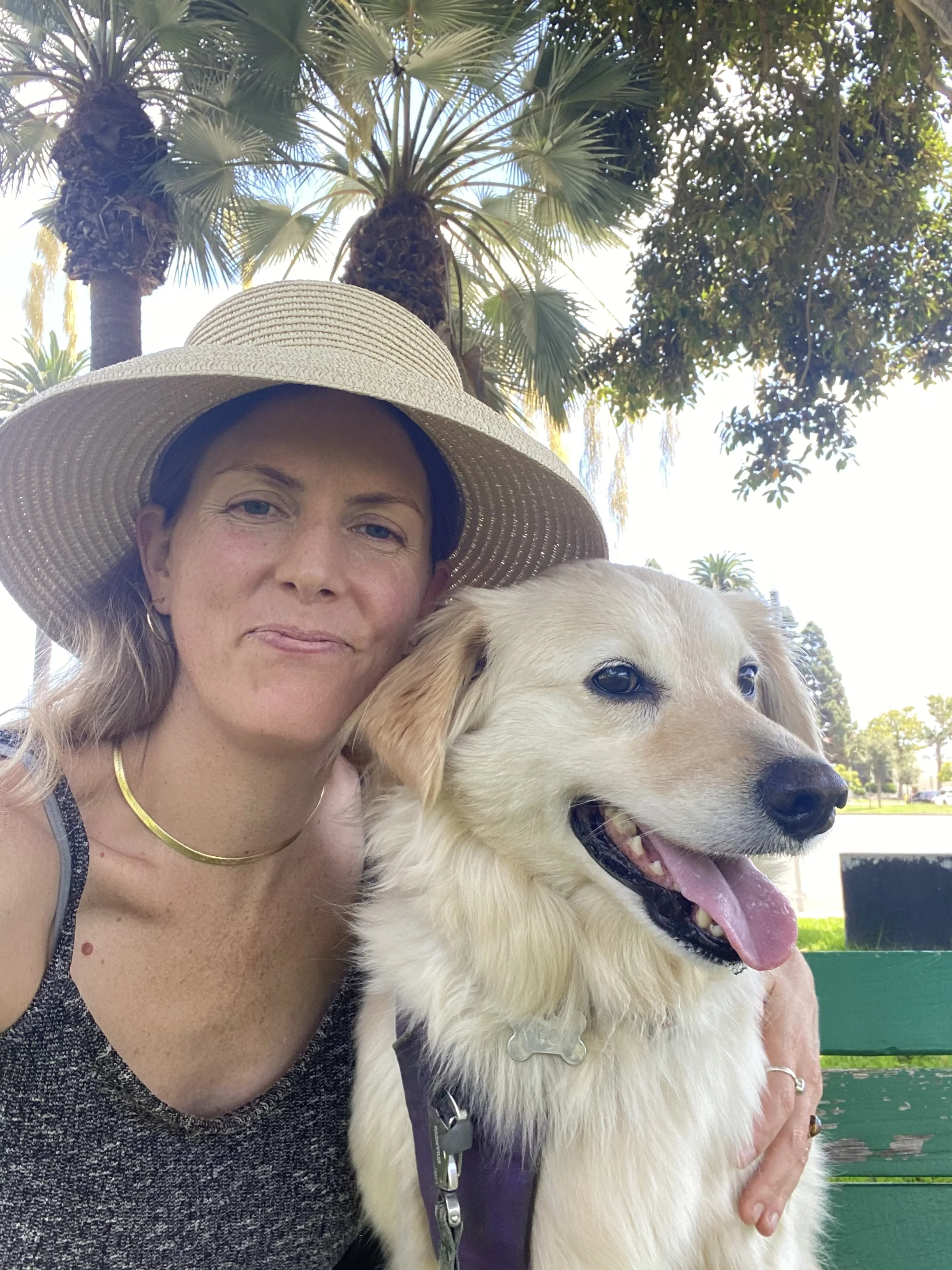 A woman wearing a large sunhat and a sleeveless top, smiling with her golden retriever dog, outdoors in a park with palm trees and clear sky in the background.