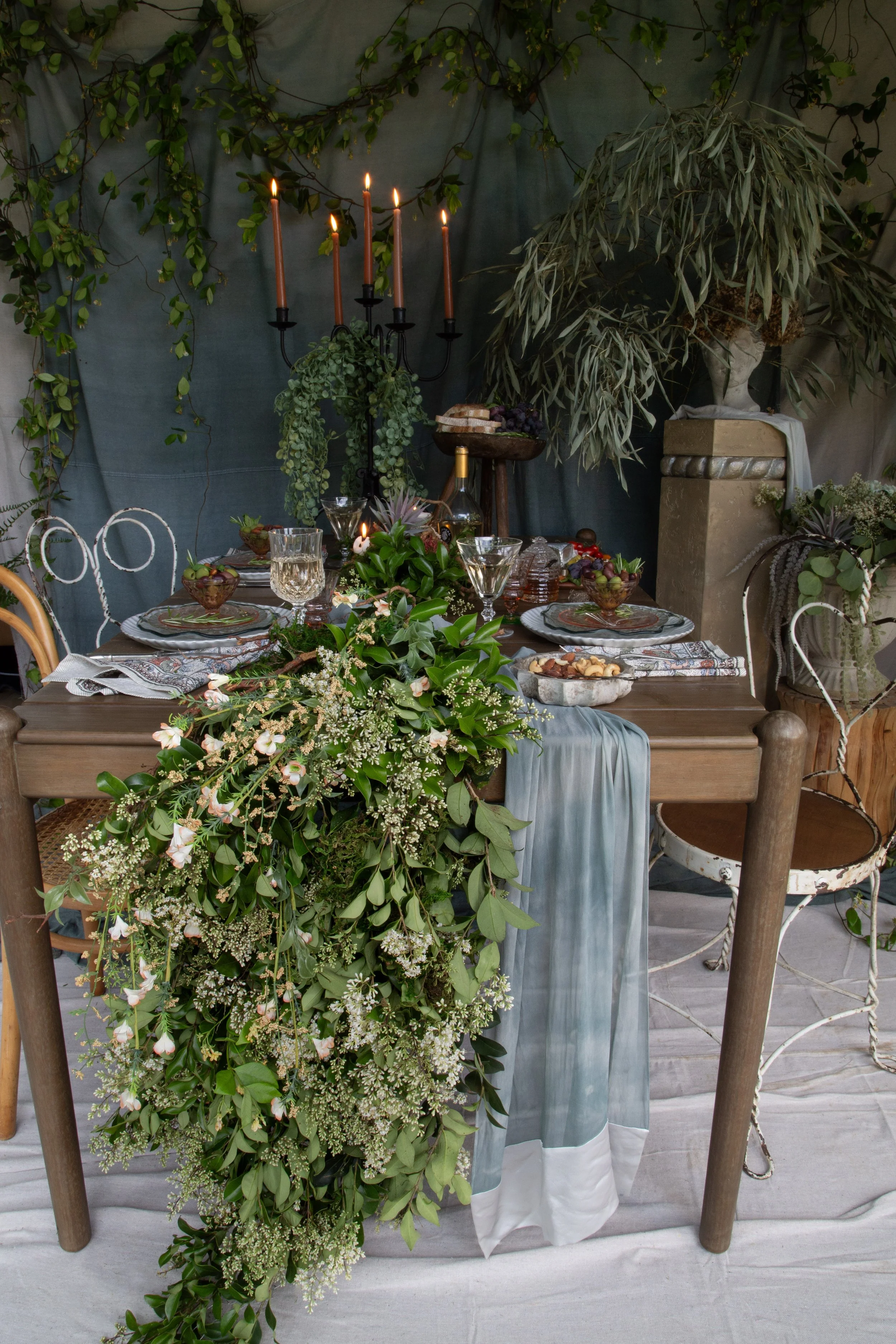 A dinner table set for a celebration with green and white floral arrangements, candles, glassware, and plates, surrounded by greenery and potted plants.