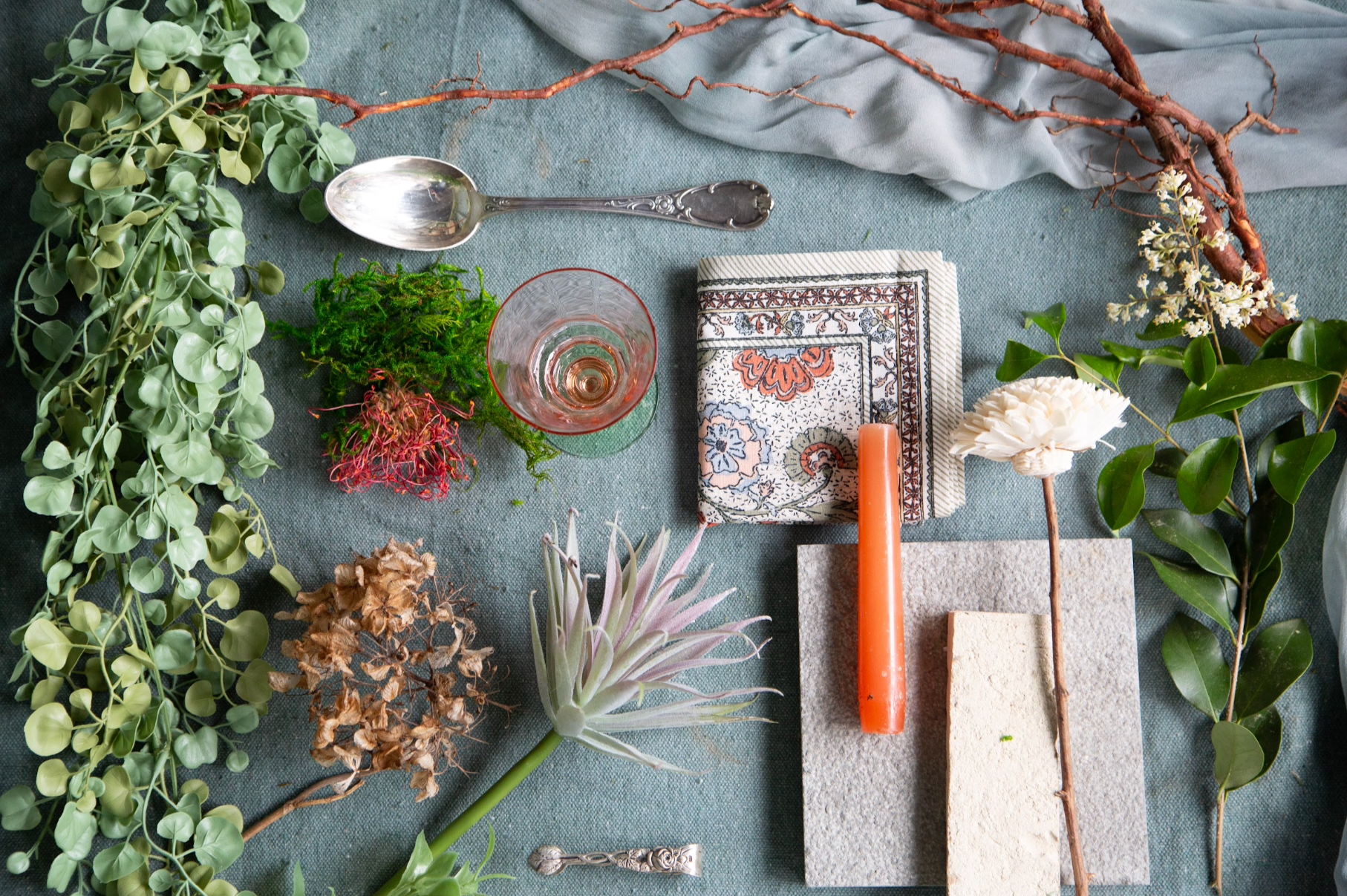 Arrangement of various plants, leaves, a candle, a glass, and decorative items on a textured fabric surface.