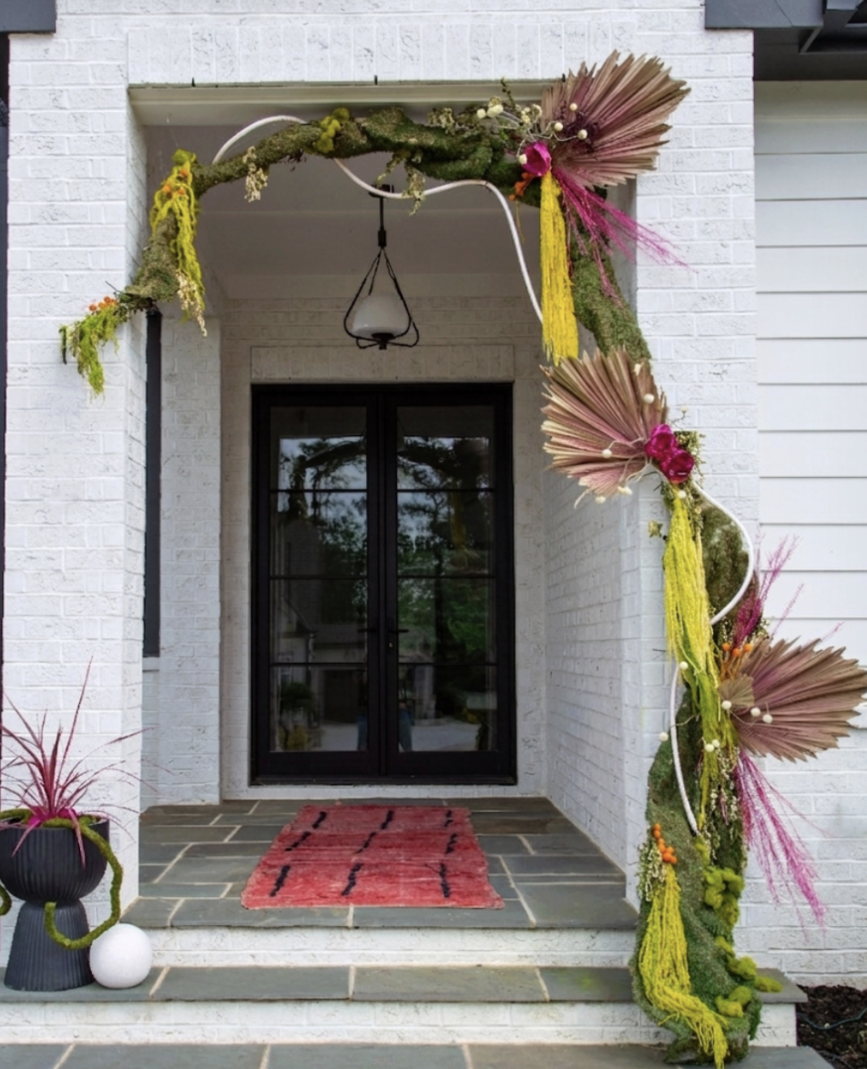 Decorative floral arrangement framing a front porch entrance with a black door, a red rug, and a black planter with pink plants.