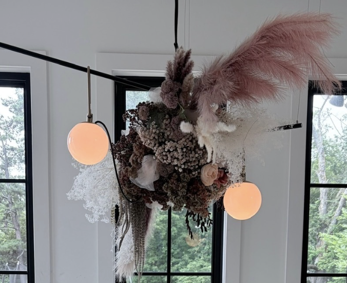 Hanging floral and pampas grass arrangement with two spherical light fixtures in front of windows.