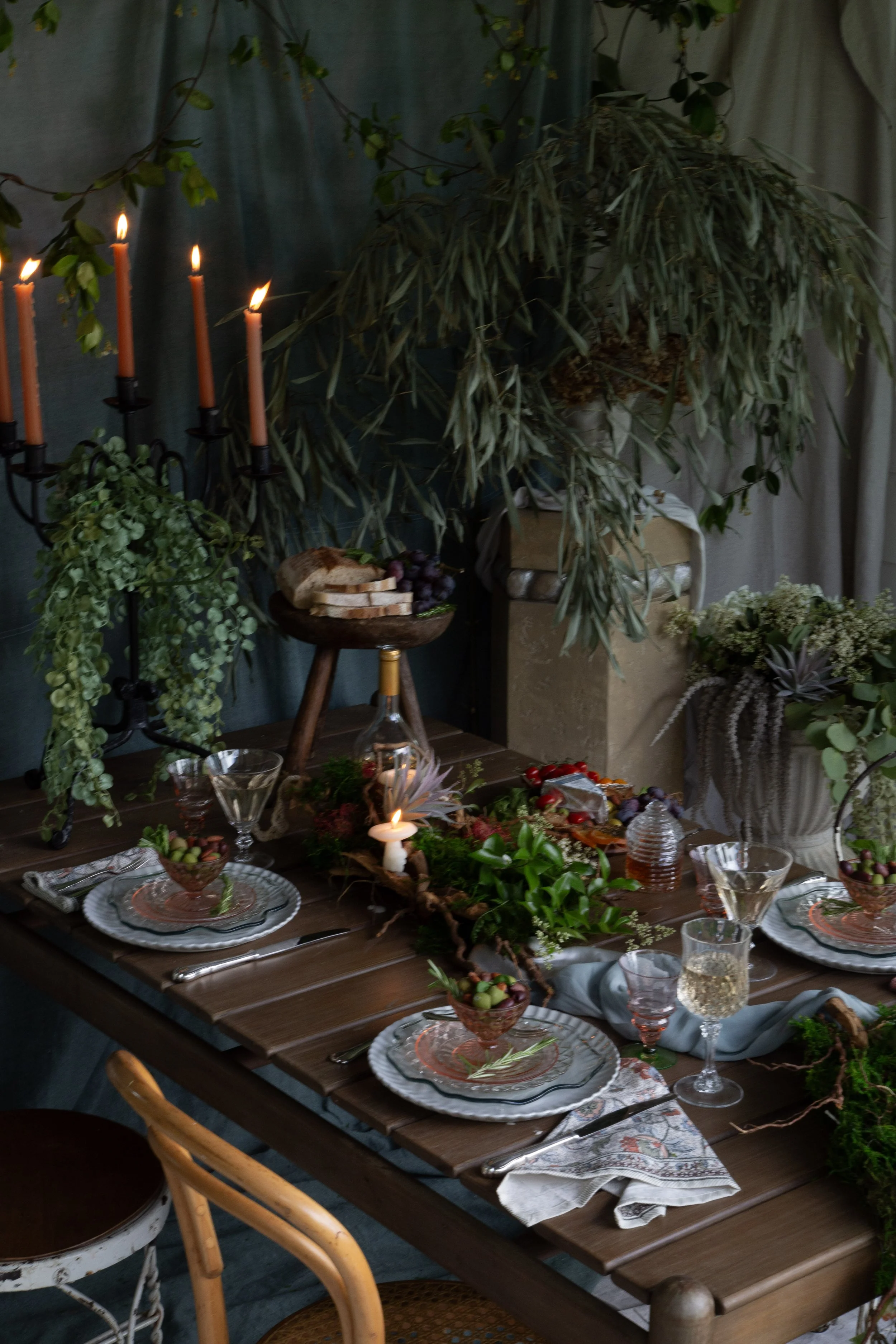 A rustic dining table decorated for a gathering with plates, glasses, and a central floral arrangement. The background features greenery, candles, and draped curtains.