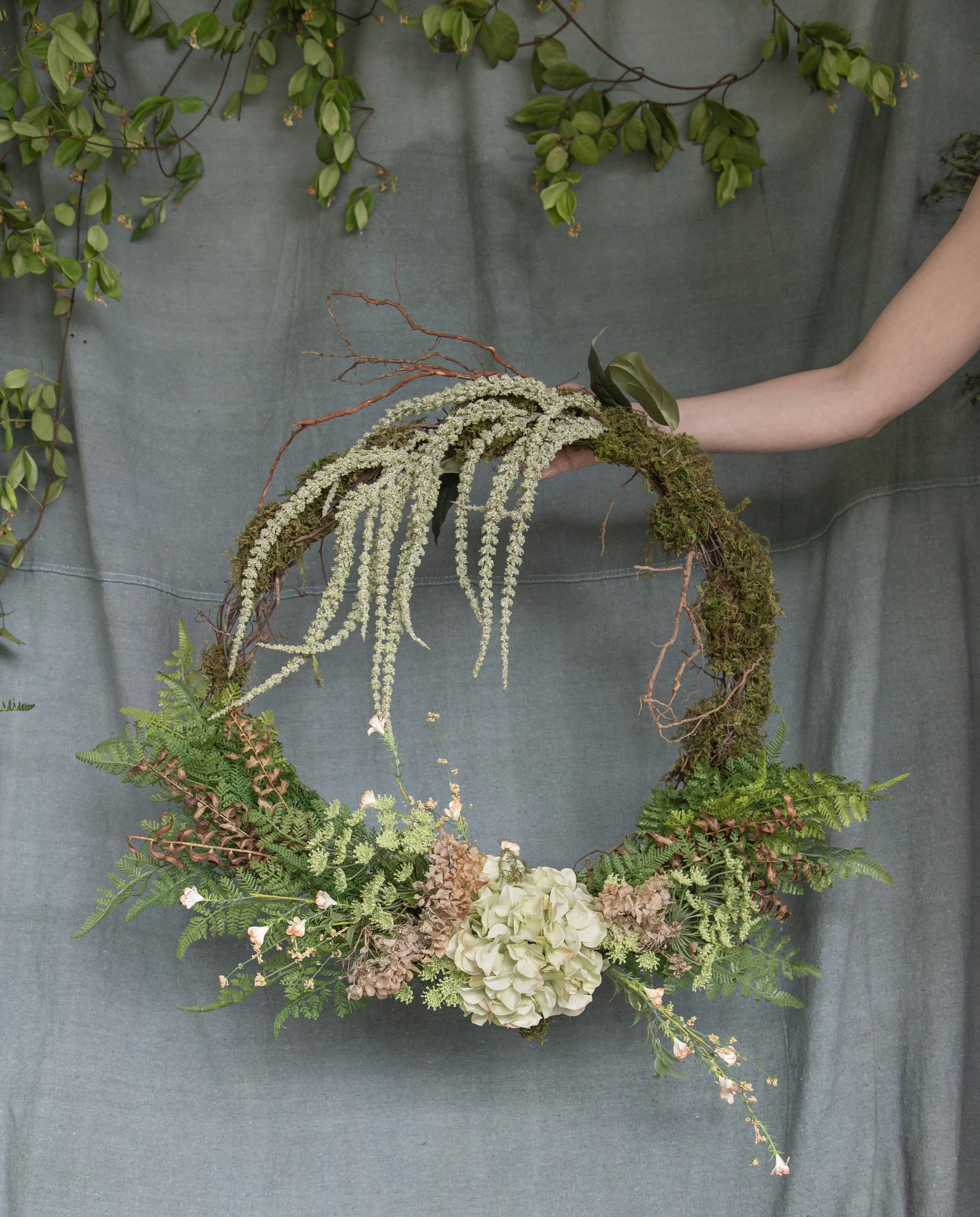A floral arrangement featuring a circular wreath with various green foliage, pale pink and white flowers, and cascading white and pink blossoms, against a dark green fabric backdrop.