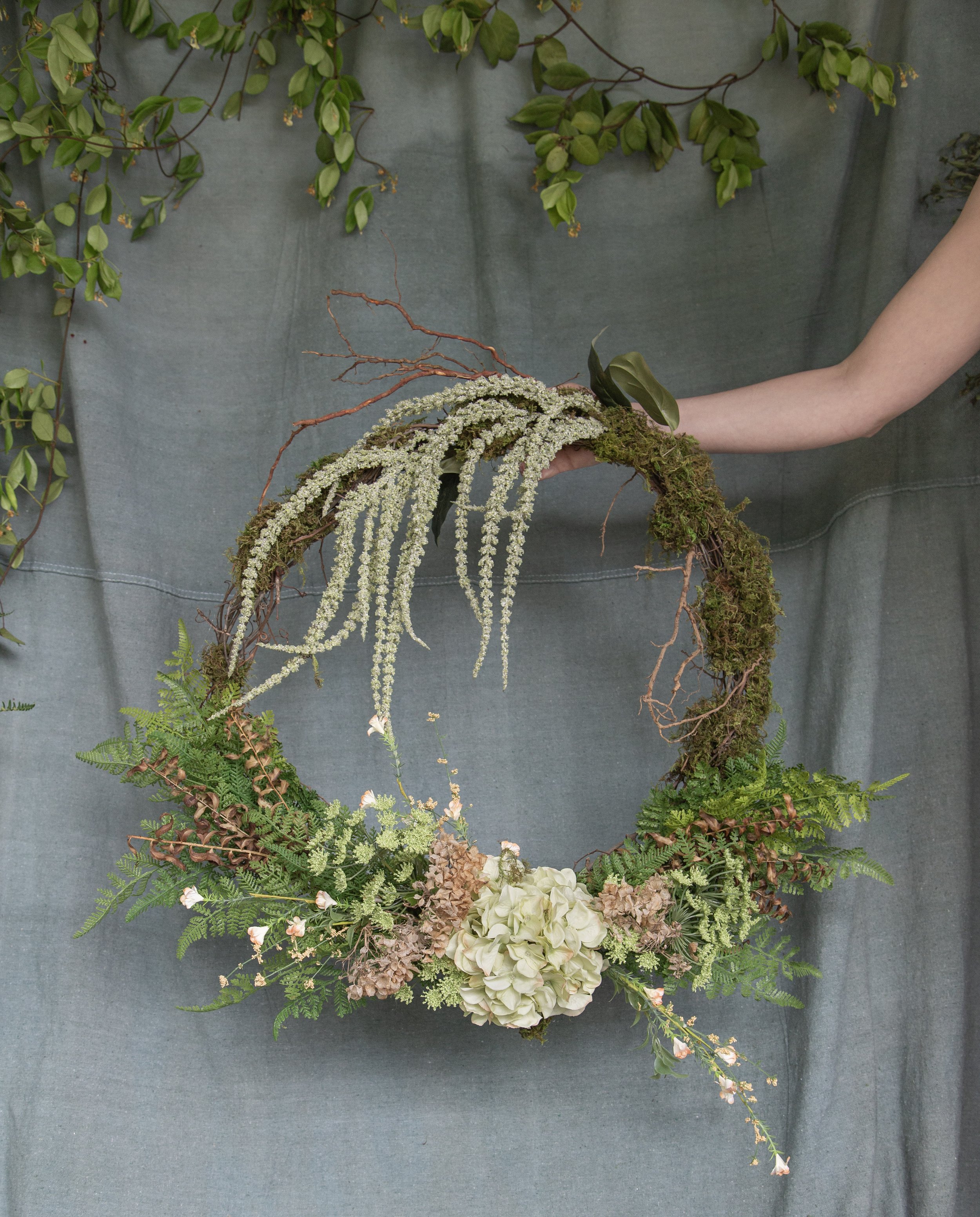 A floral arrangement featuring various green plants, ferns, moss, white cascading flowers, and dried brown flowers, displayed on a gray background.