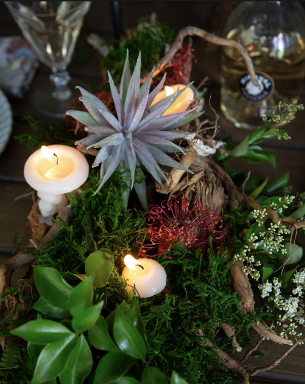 Centered arrangement of greenery, succulents, small white candles, moss, and flowers on a dark wooden surface, with glass jars in the background.