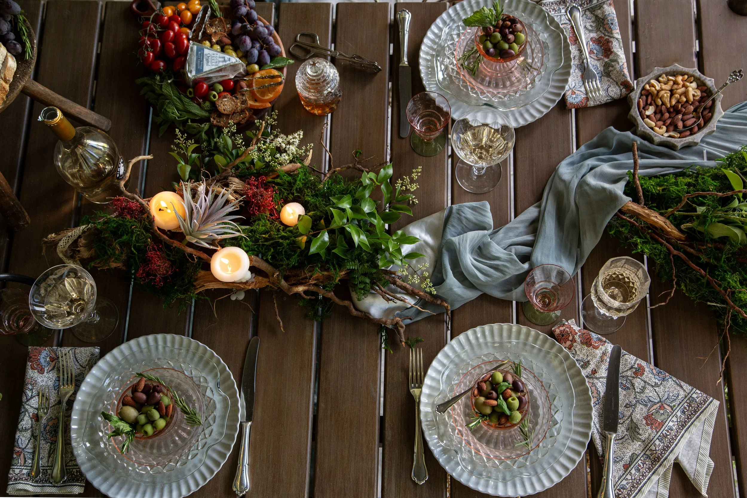 A beautifully set dining table with glass plates, gold cutlery, and a centerpiece of candles, greenery, and flowers. There are bowls of mixed nuts and clusters of grapes, along with patterned napkins and wine glasses.