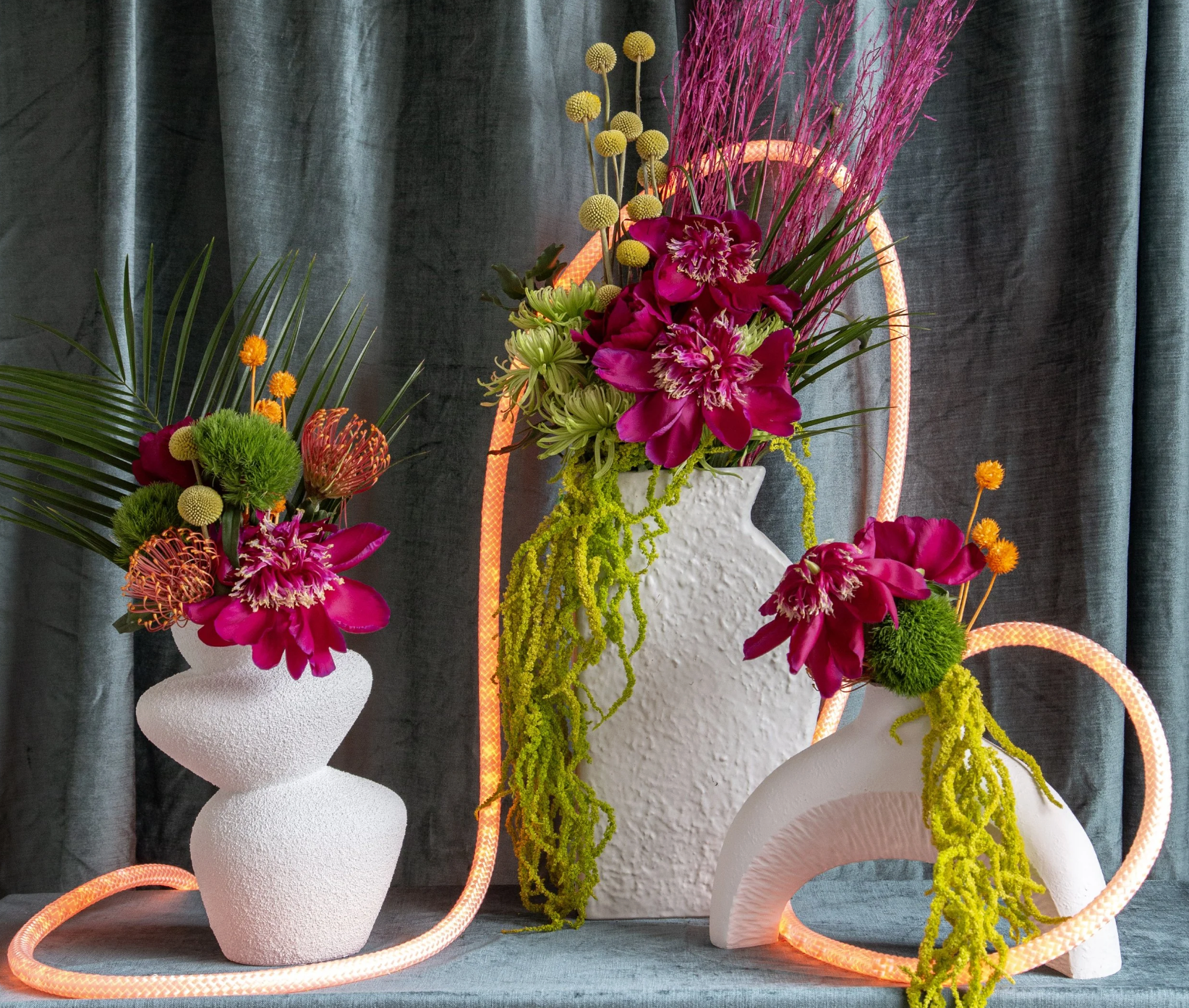 Arrangement of colorful flowers in white textured vases, with a pink illuminated circular sculpture in the background, set against dark curtains.
