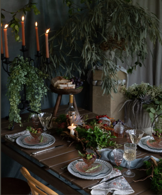 A decorated dining table with candlesticks, greenery, wine glasses, plates, and a centerpiece, set for a meal in a cozy indoor space with plants and warm lighting.