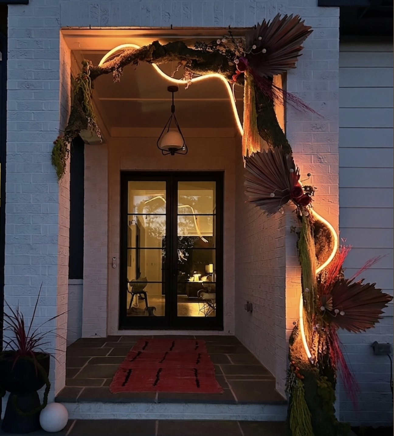 Decorated front porch at dusk with dried palm leaves and plants, illuminated by warm orange and yellow lighting, featuring a black-framed glass door and a red striped rug on stone tiled steps.