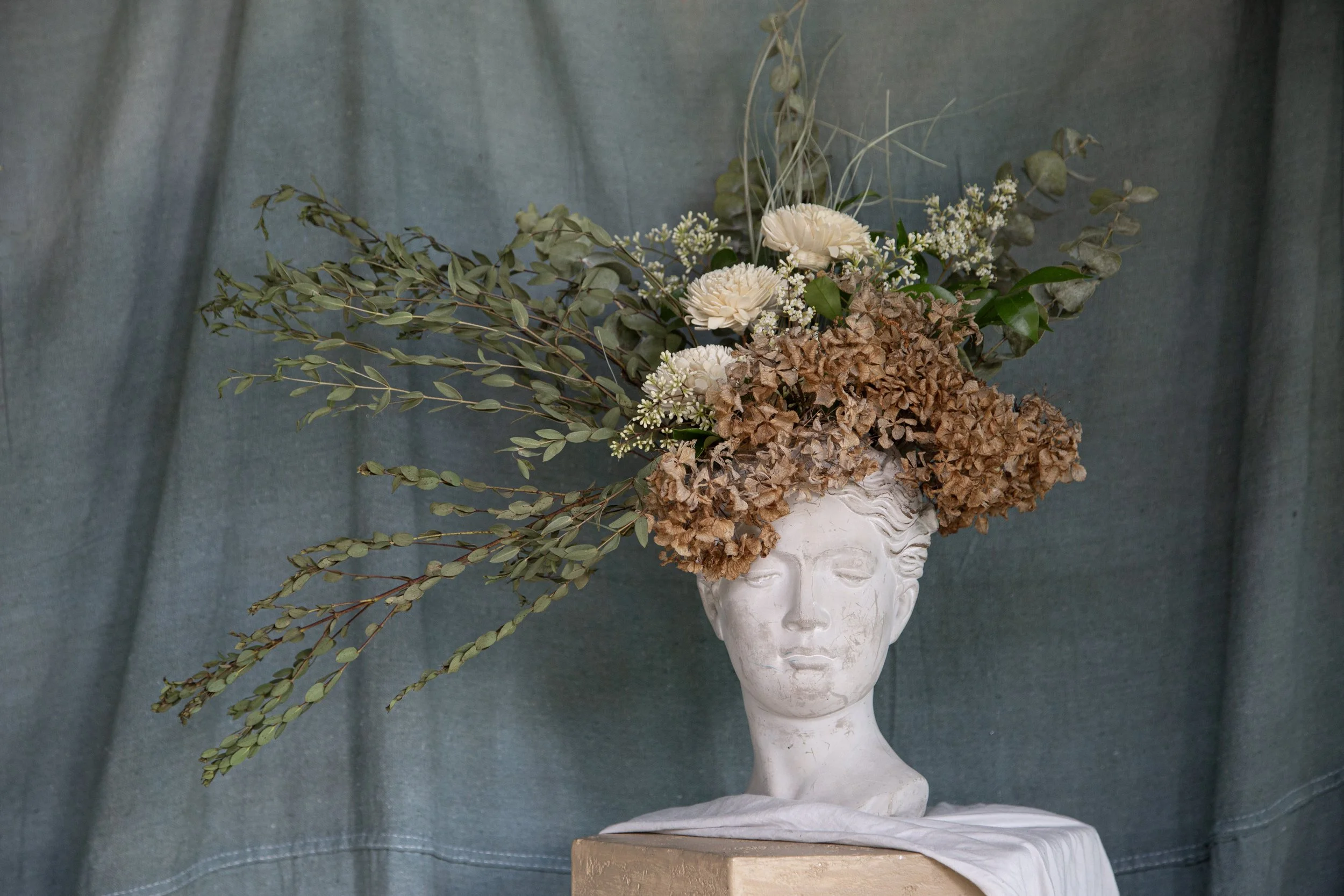 A white bust sculpture of a woman's head serving as a vase for a bouquet of dried and fresh flowers, including eucalyptus and white blossoms, against a blue fabric backdrop.