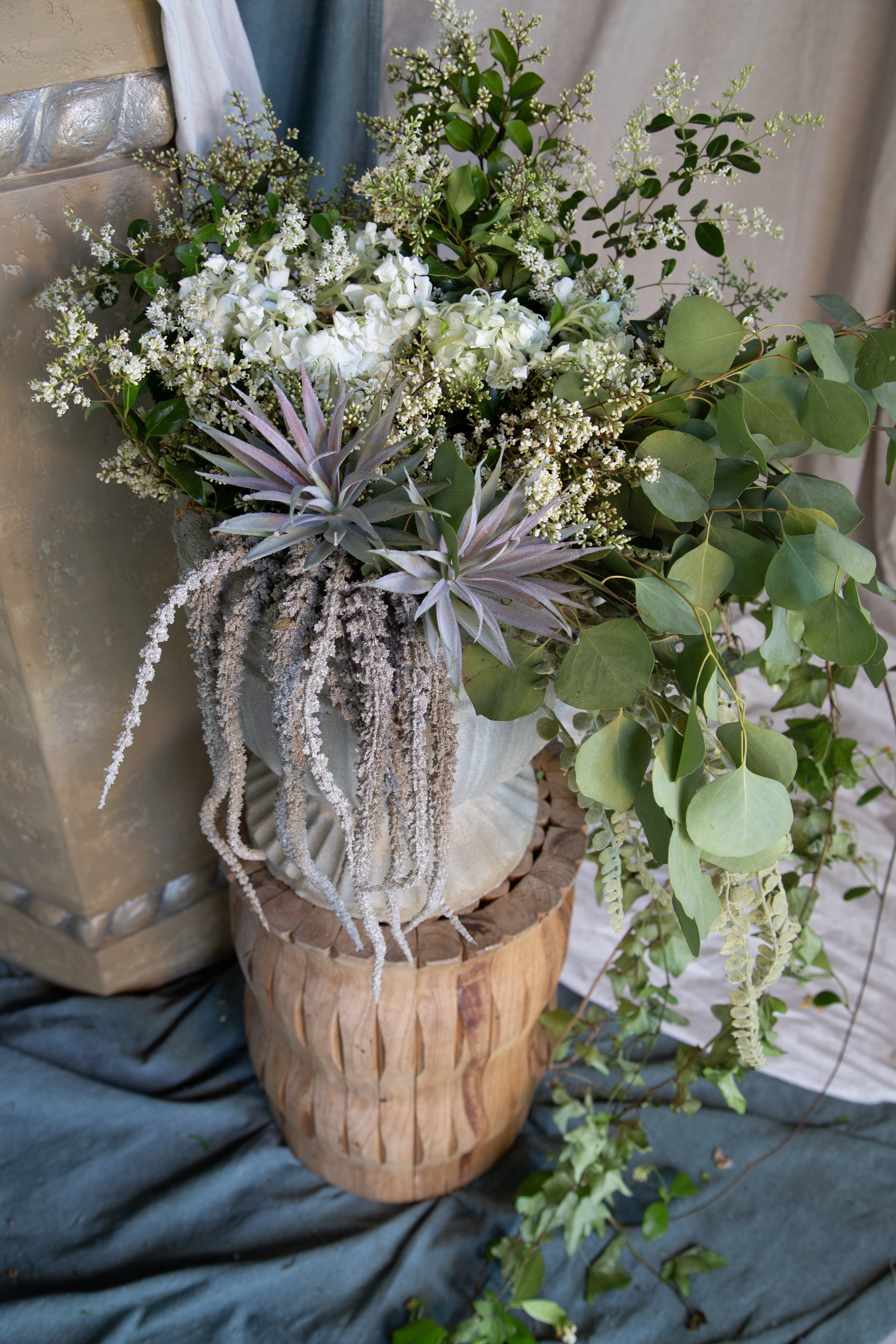 A floral arrangement consisting of white flowers, green leaves, and purple-tinted succulents in a textured, cream-colored vase on a wooden stand, with a brown brick wall and grey curtains in the background.