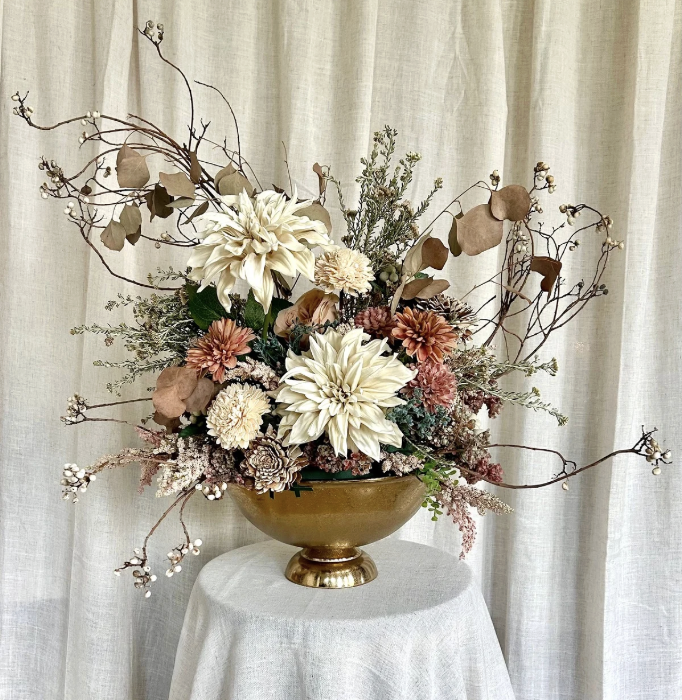 Elegant floral arrangement in a gold vase featuring large white flowers, softer pink and cream blooms, dried branches, and muted greenery, displayed on a white table with a light-colored fabric backdrop.