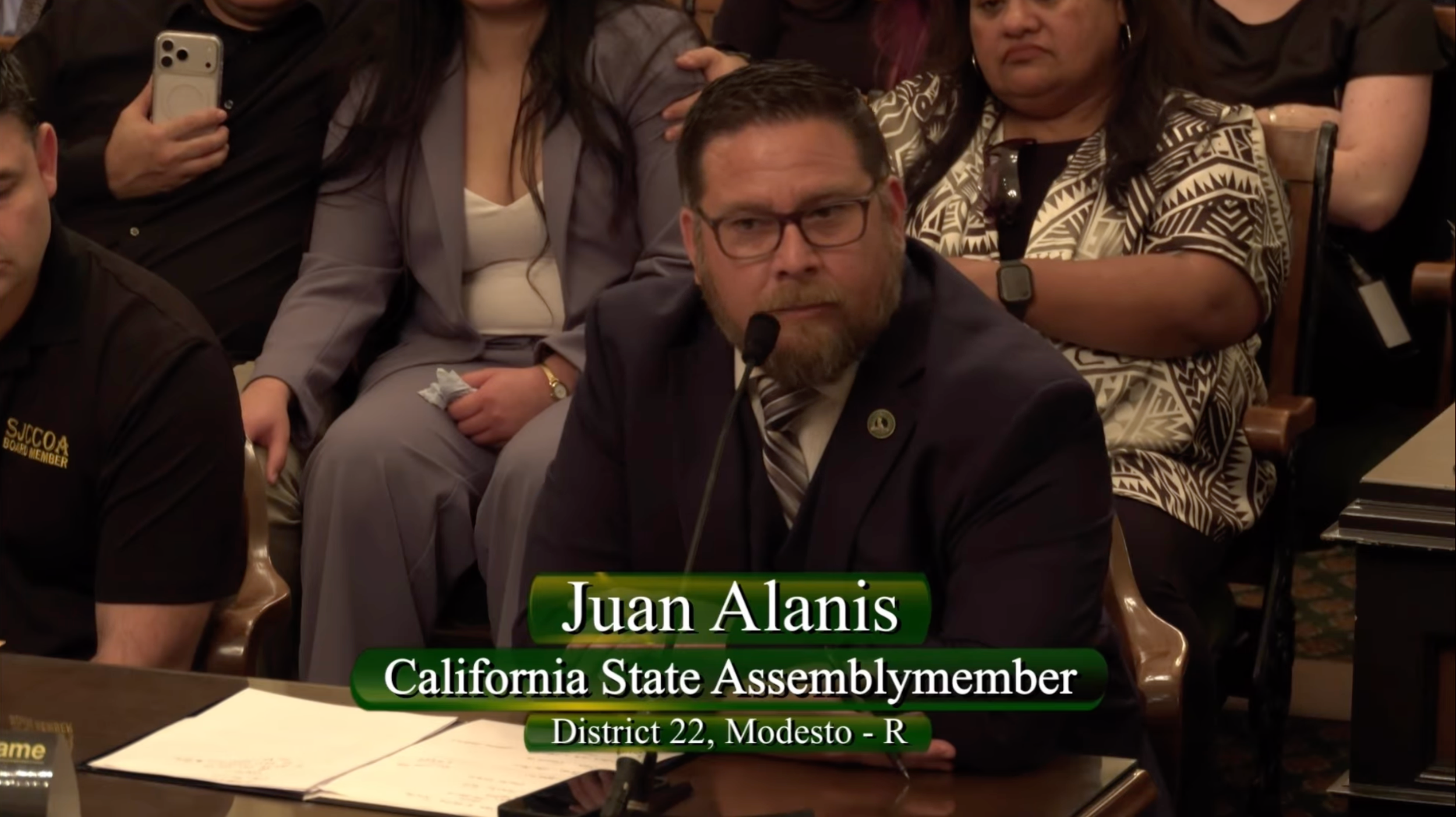 Juan Alanis, California State Assembly member from District 22, Modesto, is sitting at a table with a microphone, wearing glasses and a suit with a striped tie, in a formal meeting or hearing room with several people seated behind him.