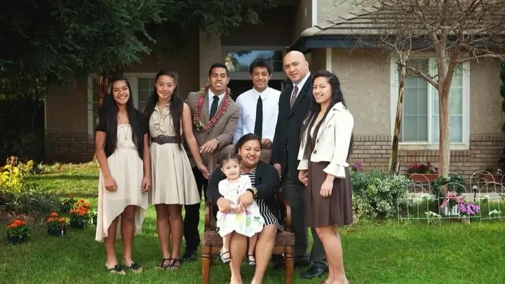 Group of seven people, including two children, standing outside in front of a house with a lawn and garden, smiling at the camera.
