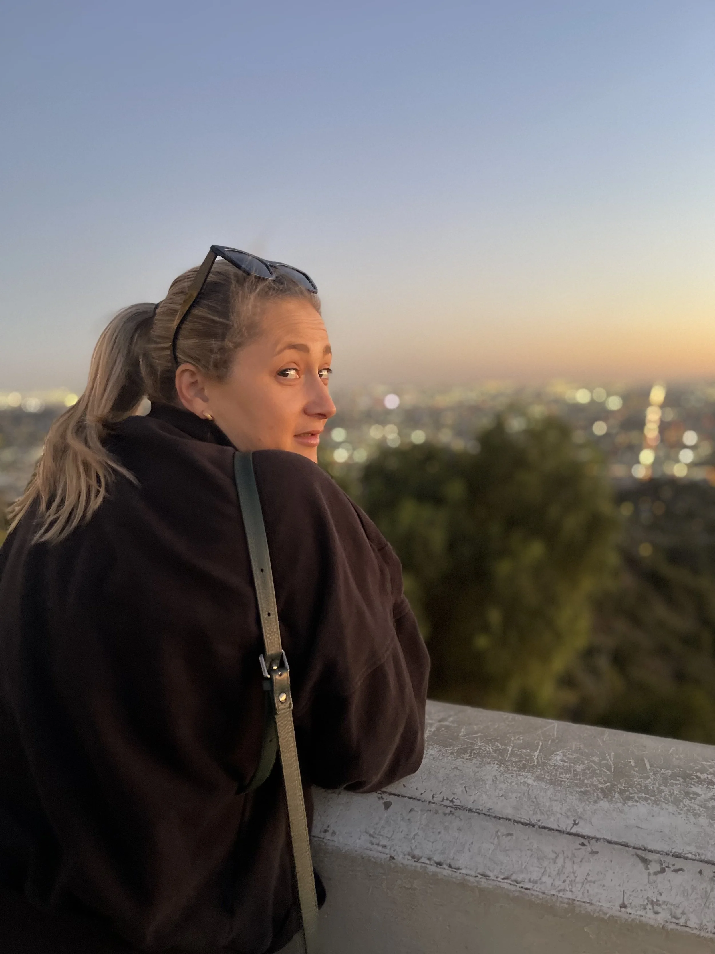 A woman with blonde hair pulled back, wearing sunglasses on her head and a dark jacket, gazes over a cityscape at sunset, with blurry city lights in the background.