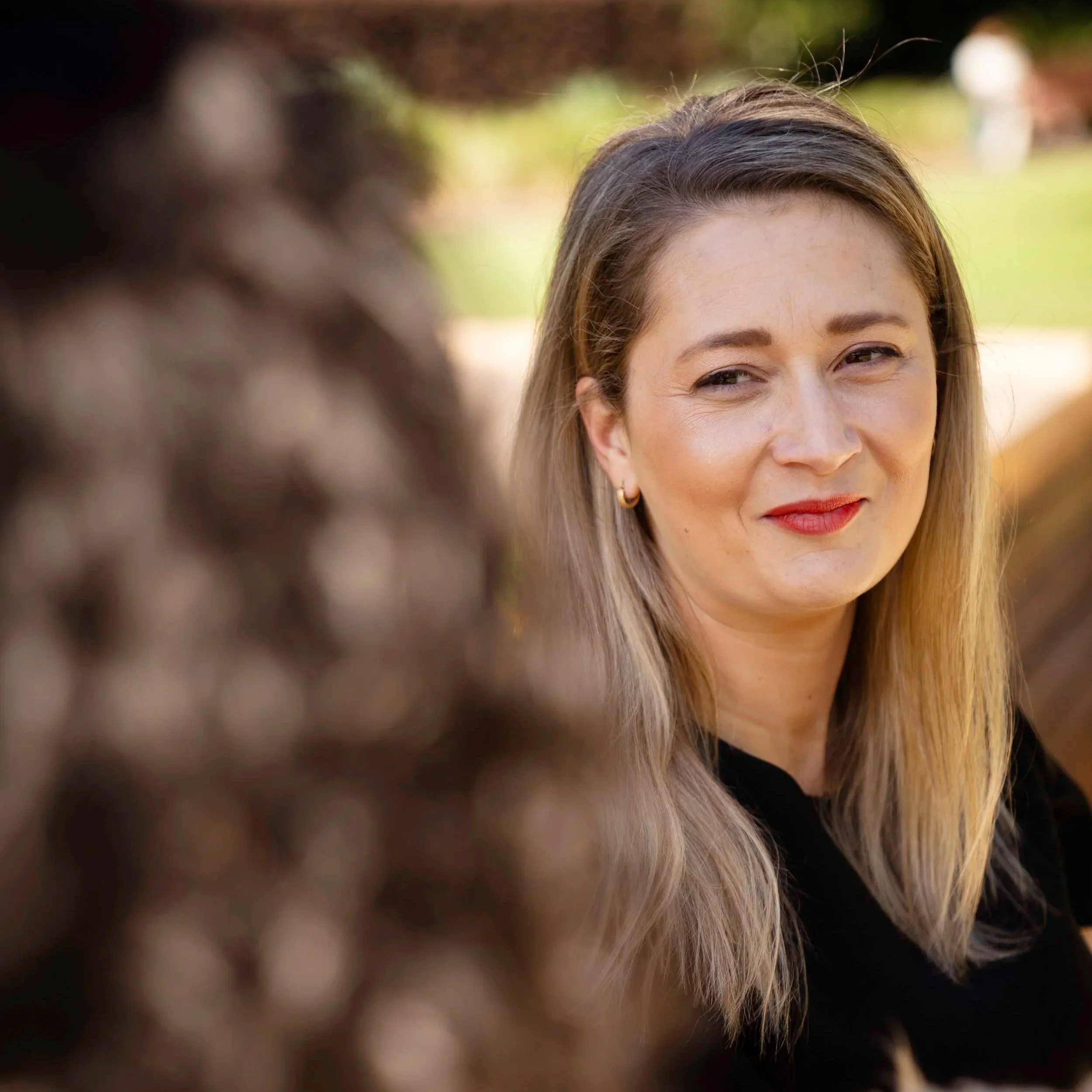 Close-up of a woman's face with light brown hair, smiling softly, outdoors with blurred green background.