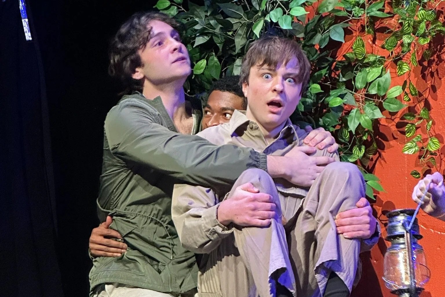 Michael Pruski, and two other men are hugging each other tightly, with one man looking off to the side and the other looking directly at the camera. The background features green foliage and an orange wall.