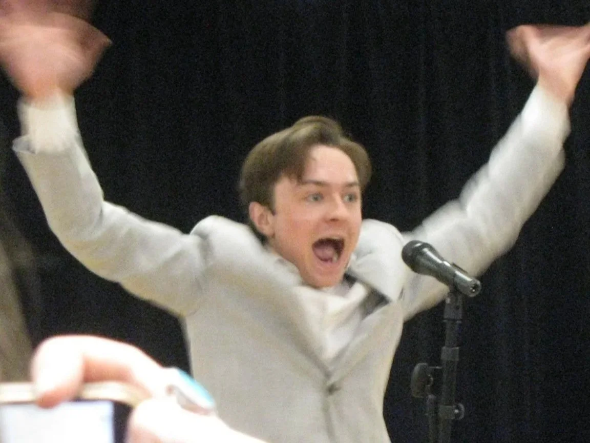 Michael Pruski dressed in a light-colored suit with a white shirt, standing in front of a black curtain, with his arms raised and mouth open in an excited and celebratory expression, speaking into a microphone. David Byrne.