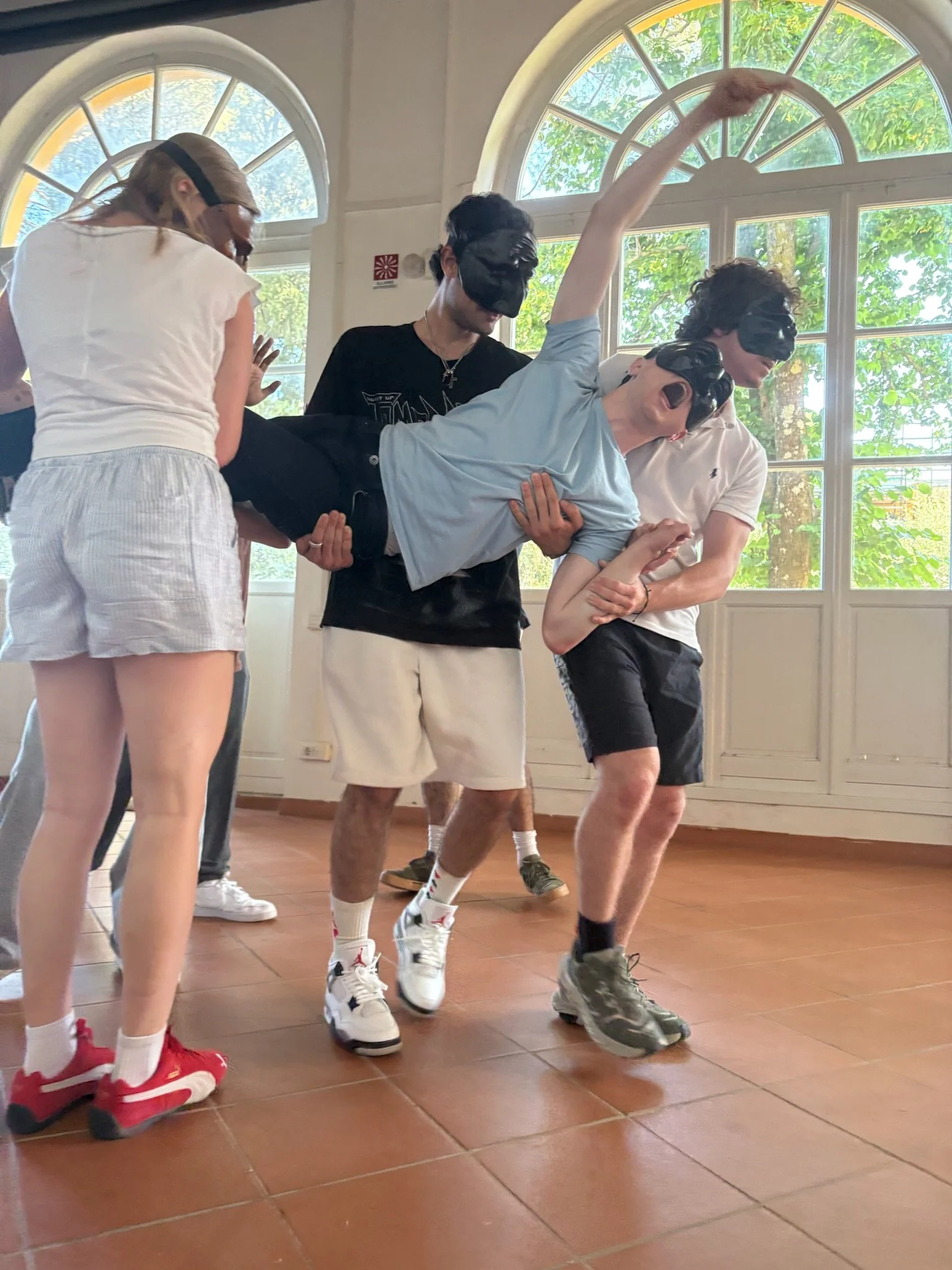Group of people wearing black Commedia dell'Arte masks, Michael Pruski, standing in a line, with one person leaning back on another person's hands.