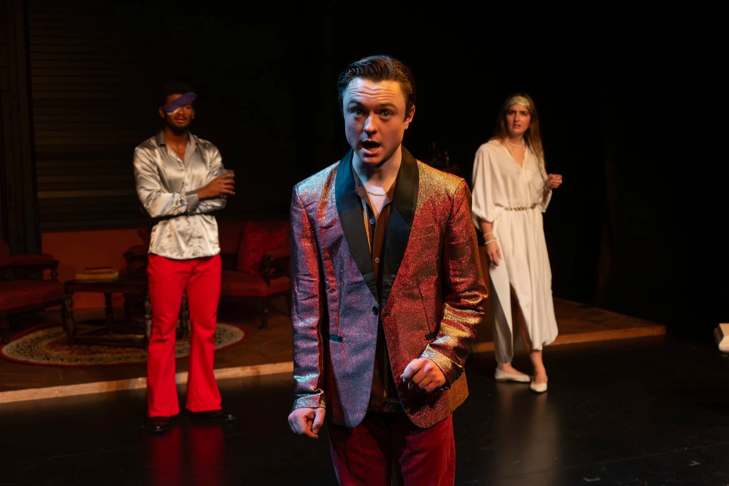 Three actors on stage in a theatrical play, with the central actor wearing a shimmering red blazer and black shirt, expressing surprise or emotion, with two other actors in the background, one in red pants and a shiny shirt, the other in a light-colored dress, under stage lighting in a dark theatrical setting.