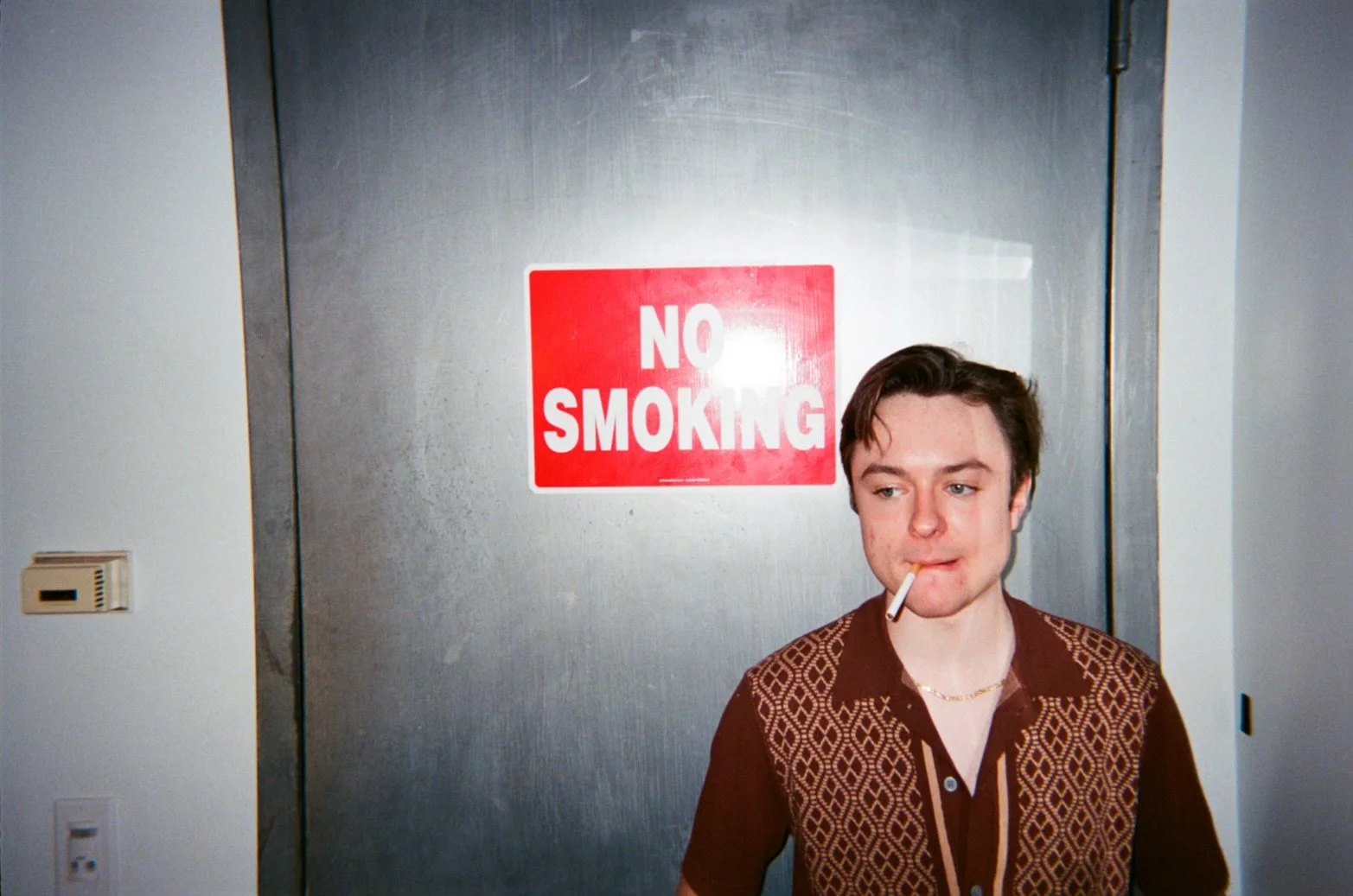 Michael Pruski with a cigarette in his mouth standing in front of a metal door with a red 'No Smoking' sign
