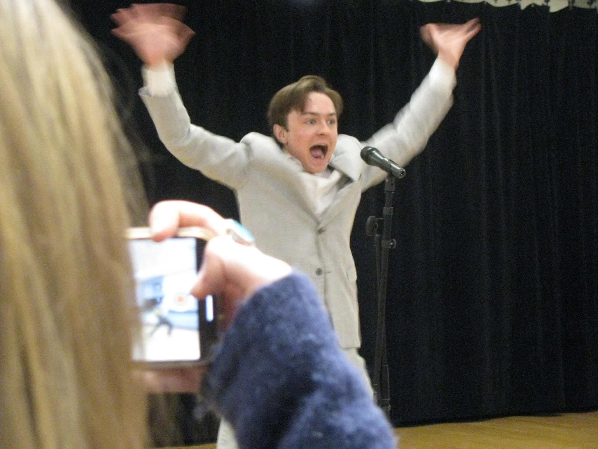 A man in a white jacket is standing behind a microphone with his arms raised and mouth open, appearing to speak passionately. He is on a stage with a black curtain backdrop. David Byrne.