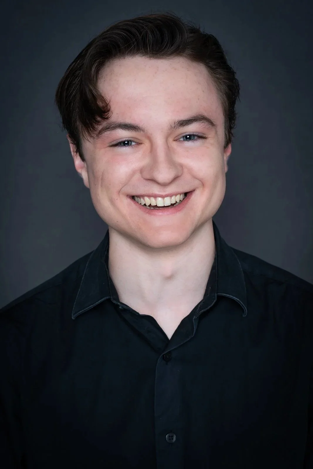 Headshot of a Michael Pruski, a young man with short brown hair, light skin, blue eyes, and a wide smile, wearing a black collared shirt against a dark gray background.