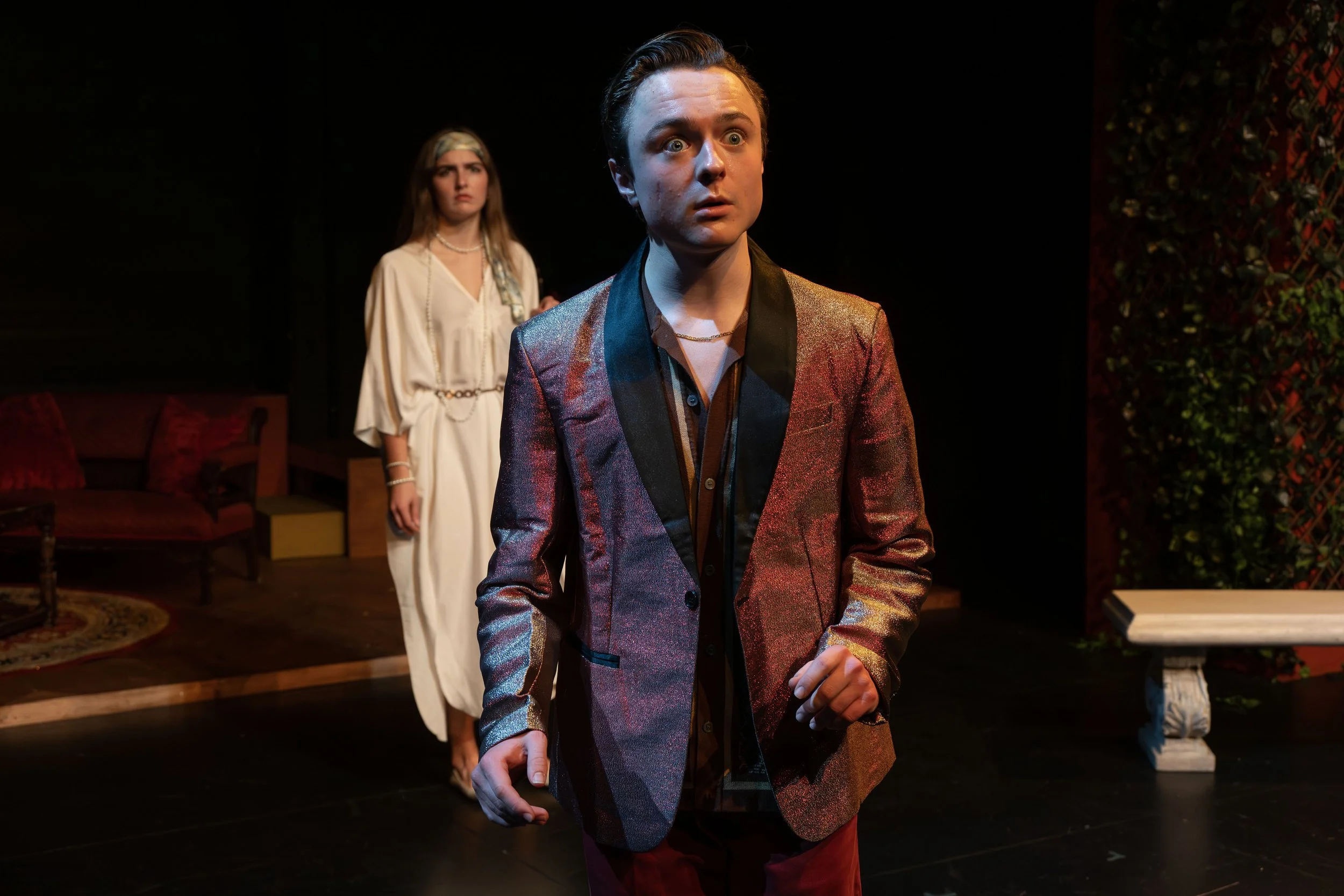 Michael Pruski in a shiny, multicolored suit with a serious expression, standing in front of a woman in a white dress with a concerned look, on a stage set with furniture and greenery.