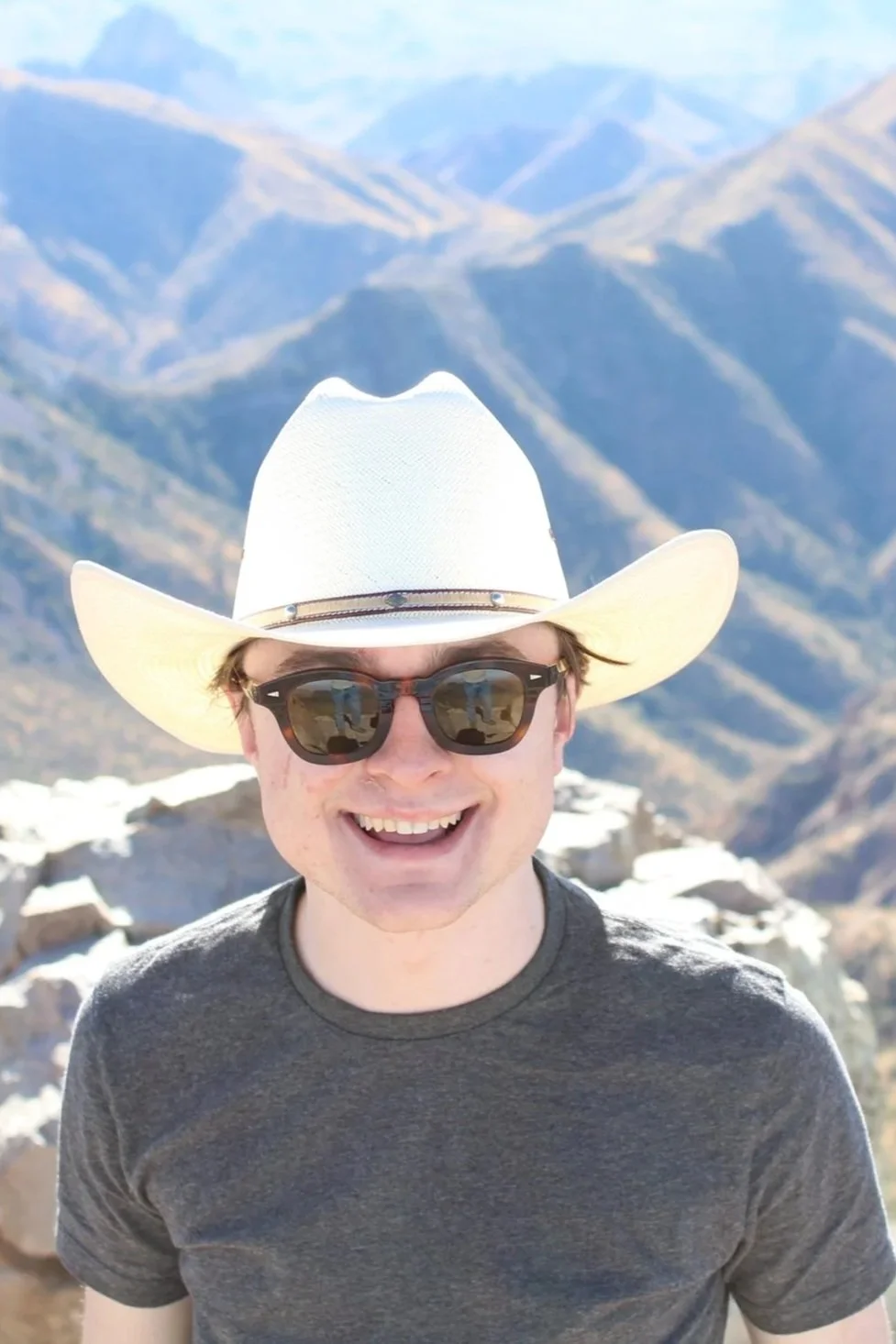 Michael Pruski wearing sunglasses and a white cowboy hat, standing outdoors in a mountainous area with rocky terrain and distant mountains in the background.