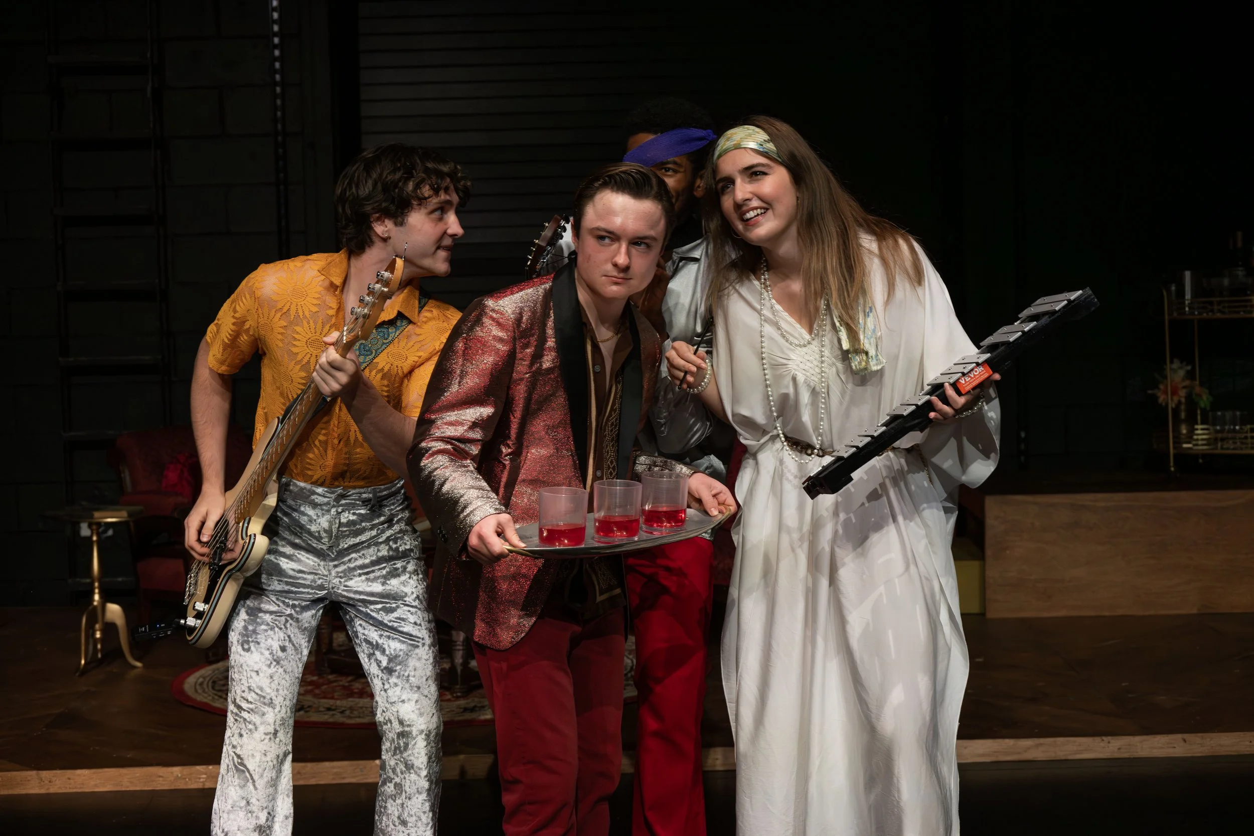 Michael Pruski and others gathered closely together on a stage, some holding musical instruments and drinks, appearing to be in a theatrical performance or rehearsal, with a dark background and some stage props.