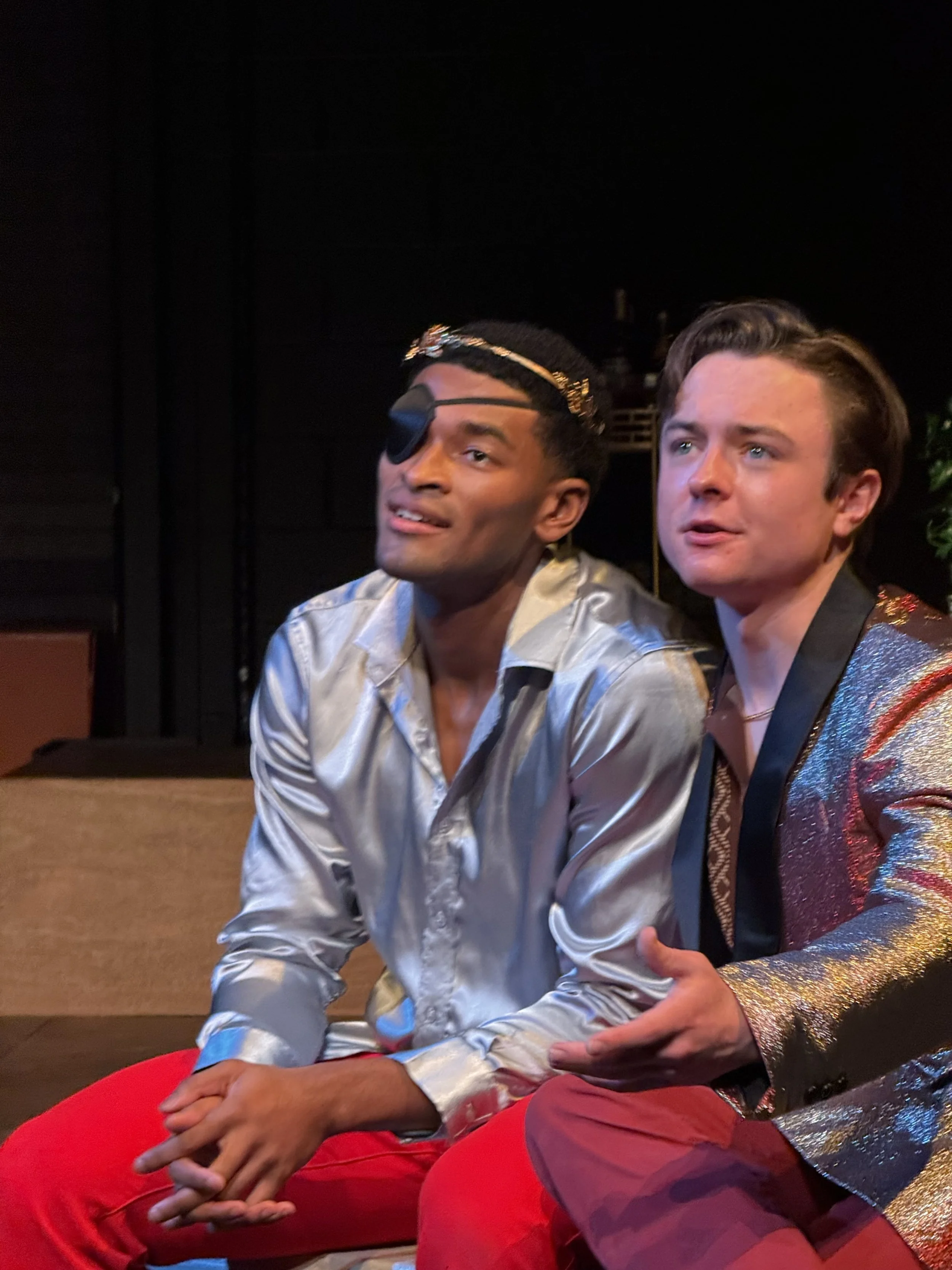Michael Pruski and man sit close together in a dimly lit setting. The man on the left has short dark hair, an eye patch over his right eye, a decorated headband, a shiny silver shirt, and red pants. The man on the right has light brown hair styled ba
