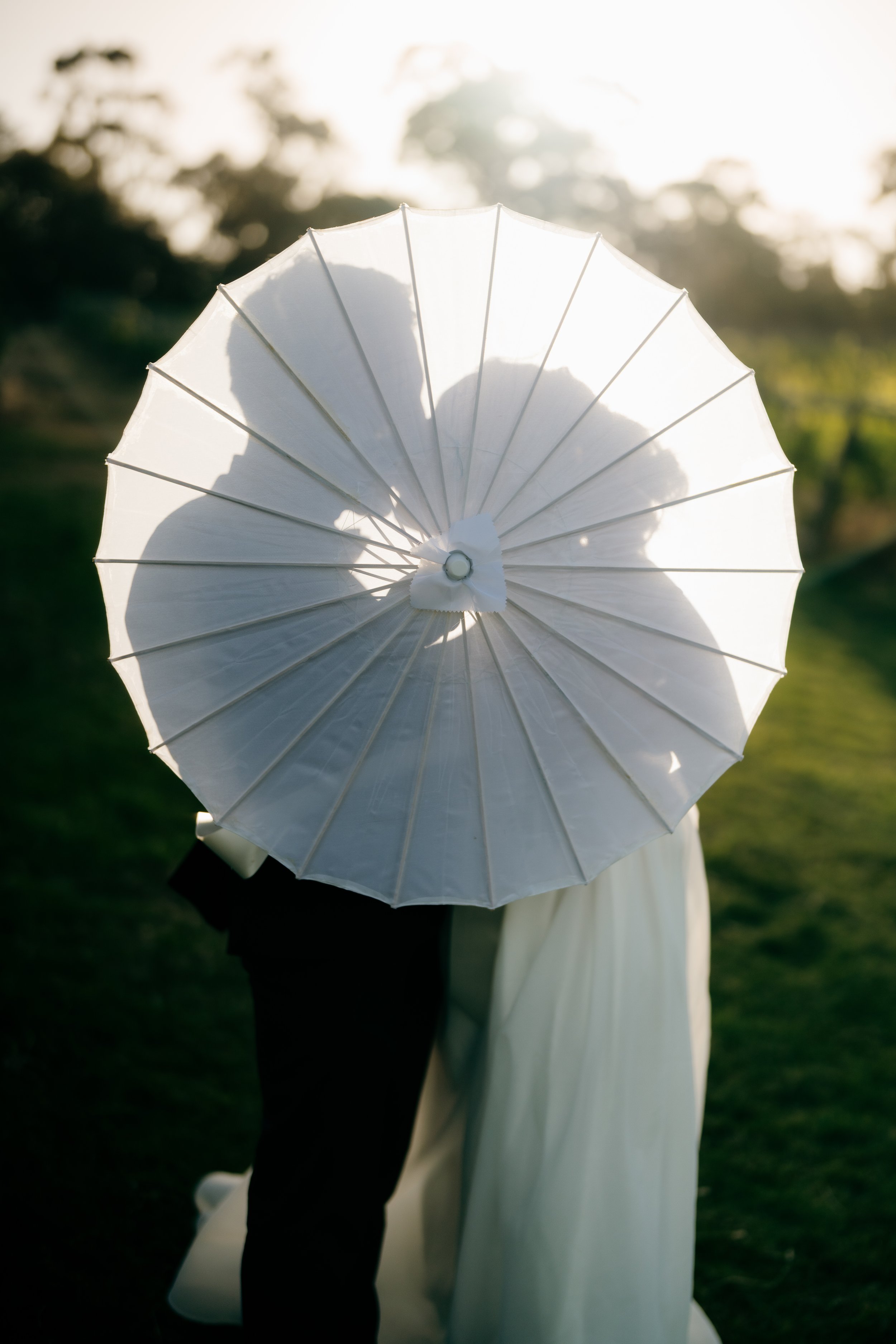 Couple standing outdoors, holding an umbrella, with the sunlight behind them creating a silhouette effect.