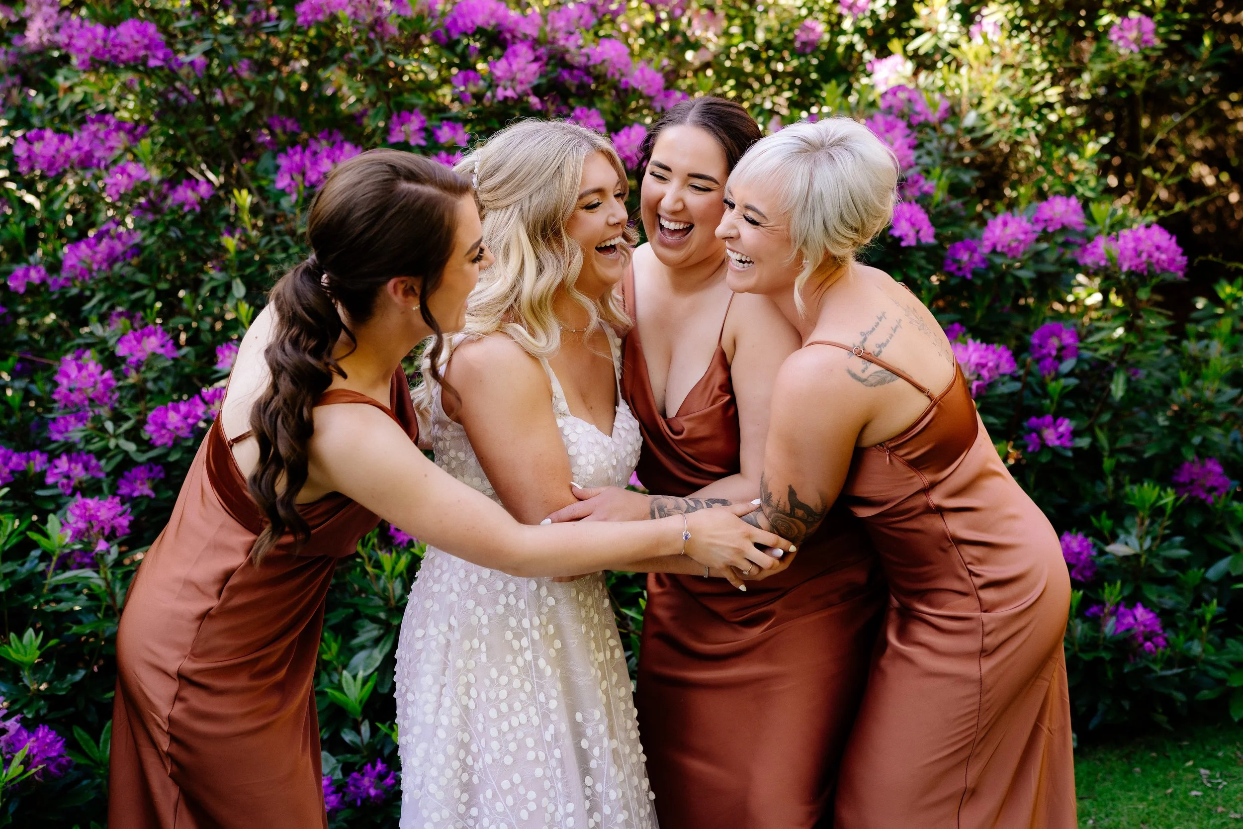 Five women are laughing and hugging in front of purple flowering bushes, dressed in casual dresses, celebrating together.