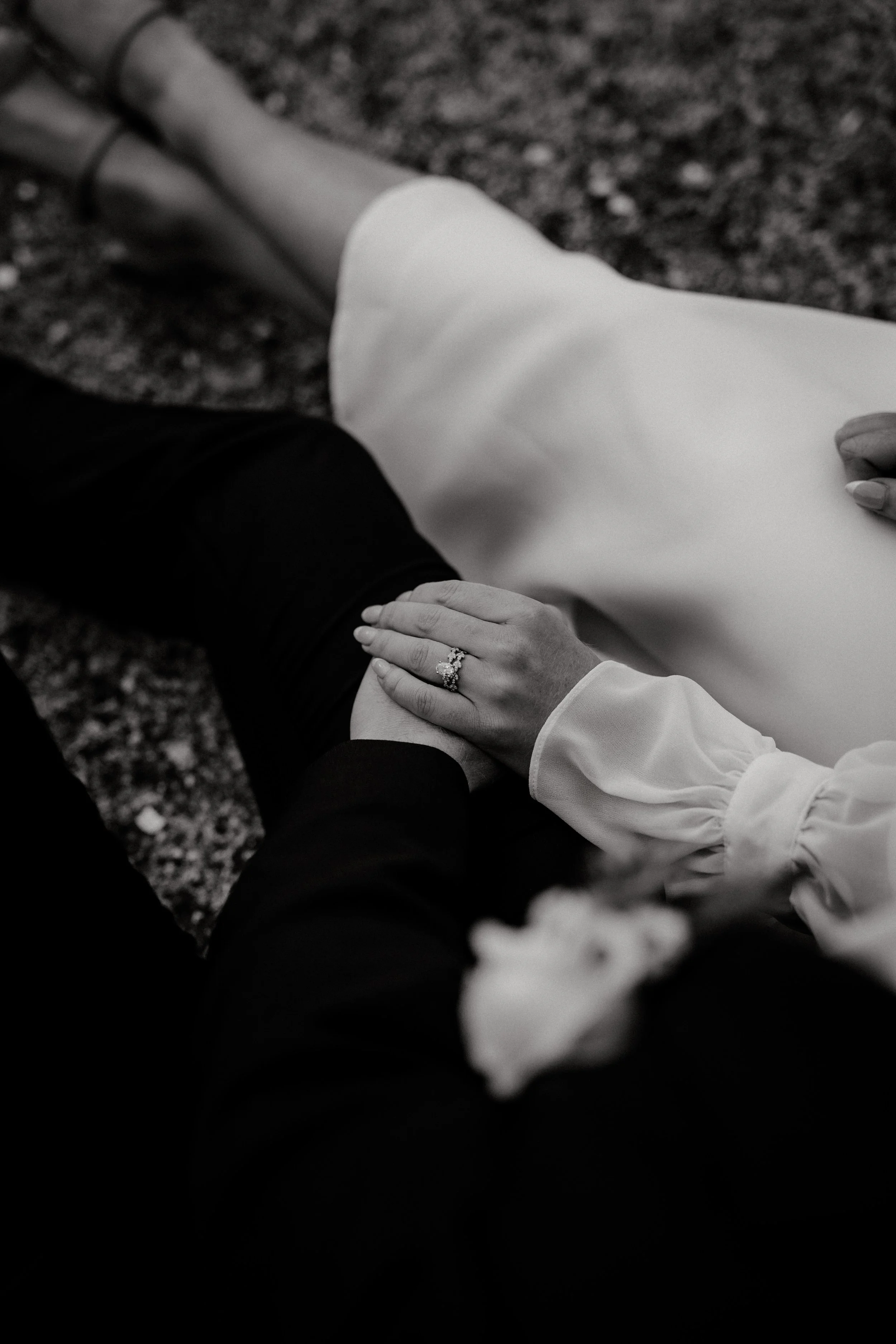 Black and white photo of a person in a wedding dress with a hand resting on their partner's knee, showing a wedding ring.