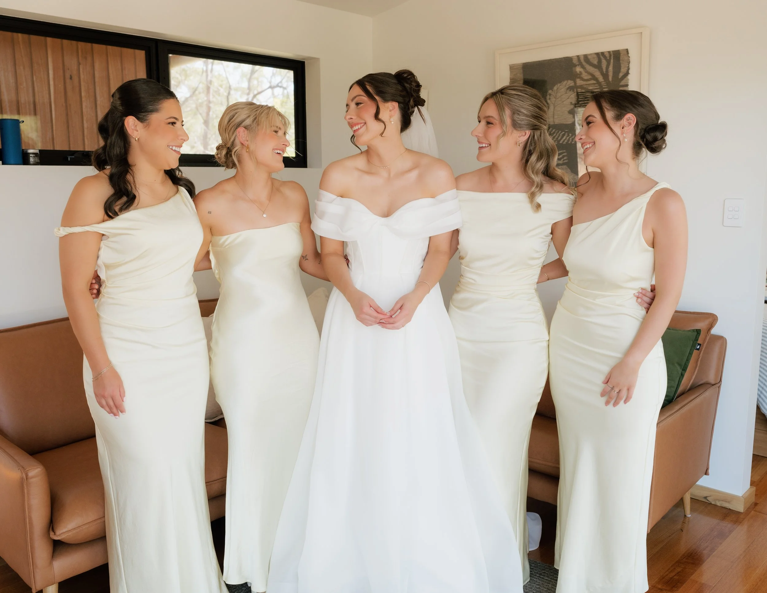 Bride in a white wedding dress with five bridesmaids in matching off-shoulder cream dresses smiling and talking in a room with modern decor.