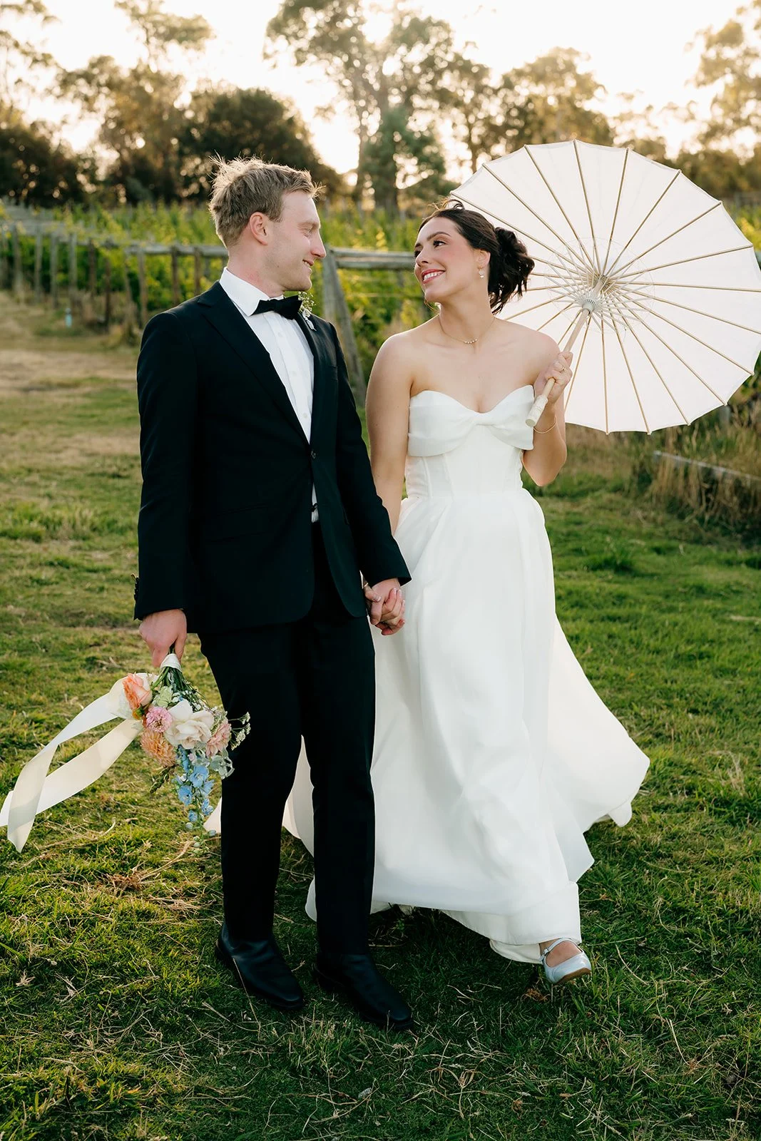 A bride and groom holding hands outdoors, with the bride holding a white parasol, both smiling at each other.