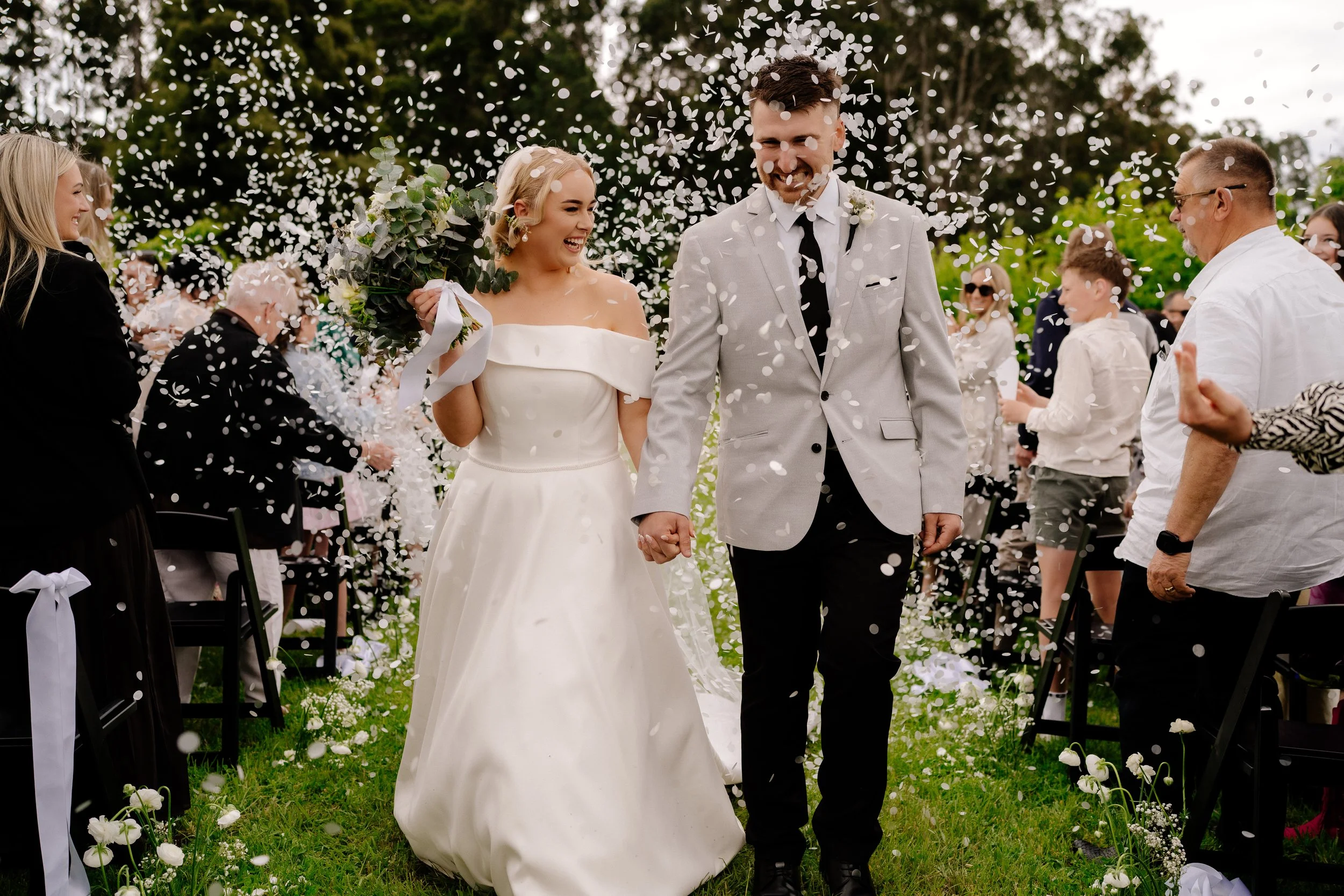 A newlywed couple walks hand in hand down the aisle at their outdoor wedding, surrounded by guests, as confetti falls around them.