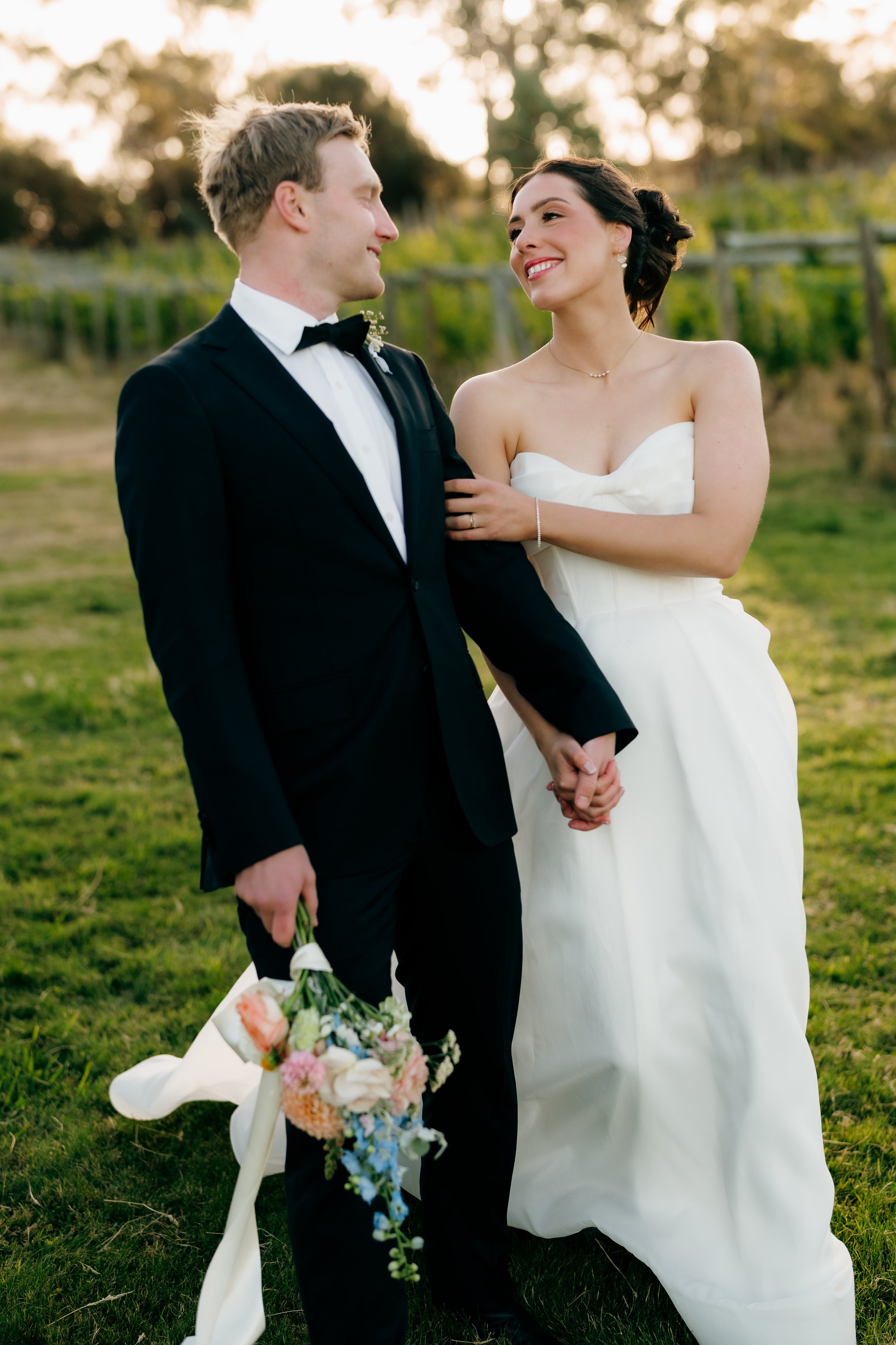 A newlywed couple holding hands outdoors during sunset, with the groom in a black tuxedo and the bride in a strapless white wedding gown, smiling at each other.