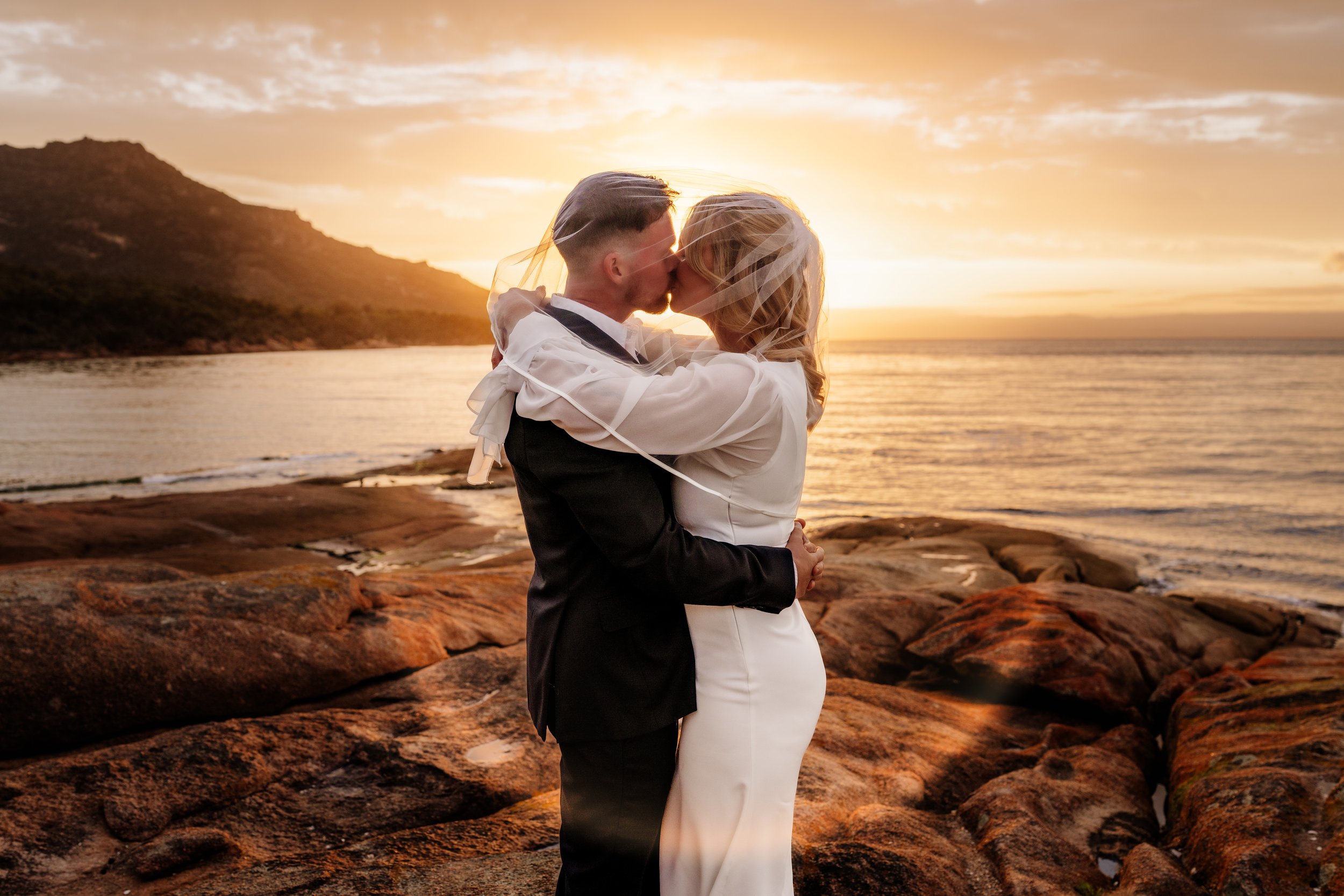 A couple dressed in wedding attire embraces and kisses on a rocky beach at sunset.