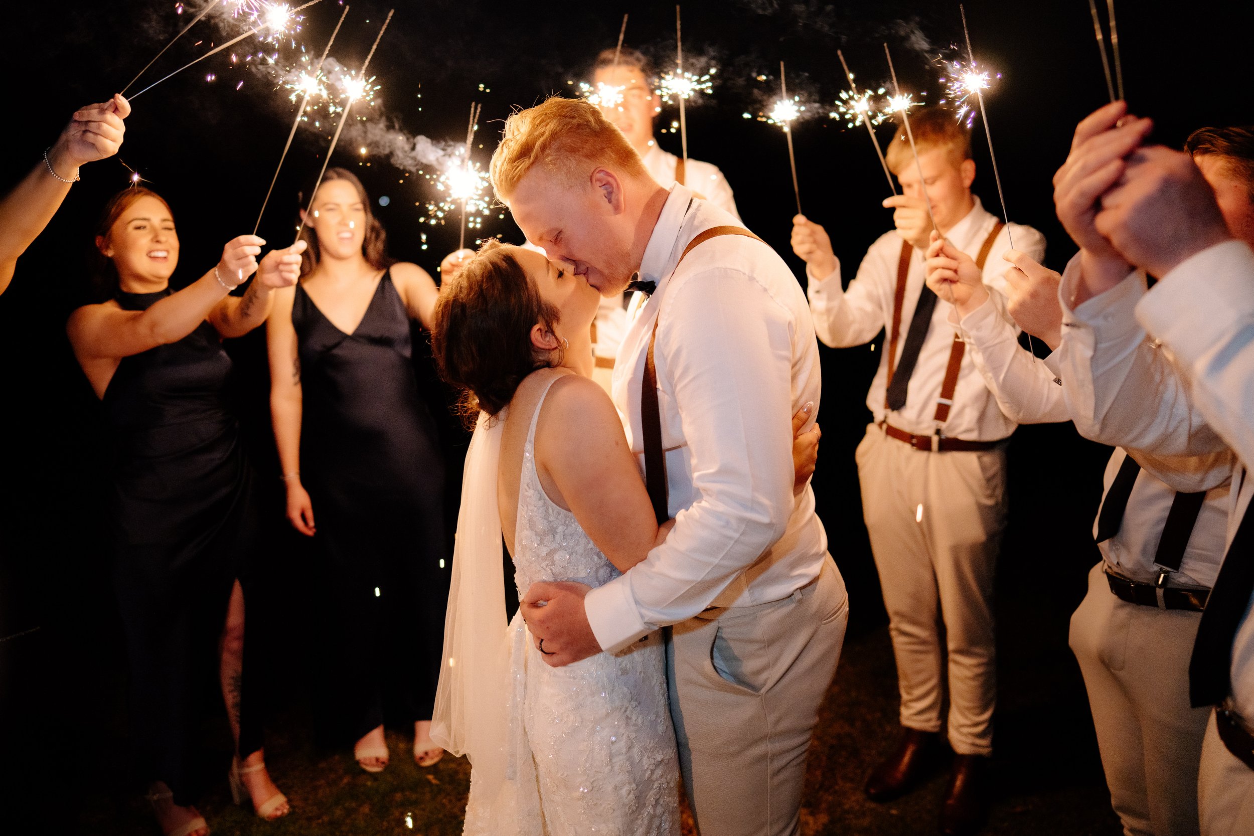 A newlywed couple sharing a kiss at night, surrounded by friends holding sparklers during a celebration.